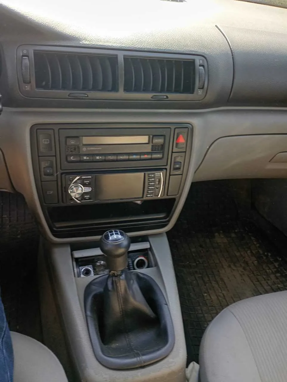 Wnętrze samochodu z widoczną deską rozdzielczą, dwoma klimatyzacyjnymi wydmuchami, dwoma urządzeniami audio (górne z przyciskami i wyświetlaczem, dolne z pokrętłem i ekranem), dźwignią zmiany biegów z czarną osłoną i oznaczeniami biegów, fragmentem jasnoszarego fotela oraz czarną podłogą z matą. W lewym dolnym rogu widoczny fragment nogi w niebieskich dżinsach.