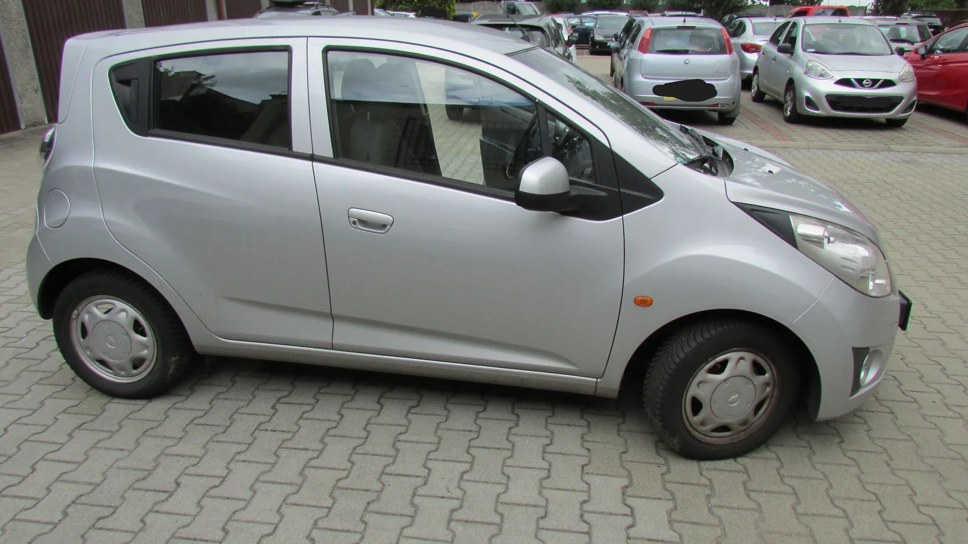 Srebrny, pięciodrzwiowy samochód miejski zaparkowany bokiem na kostce brukowej. W tle widoczne są inne zaparkowane samochody, w tym biały i srebrny model marki Nissan oraz czerwony samochód.
