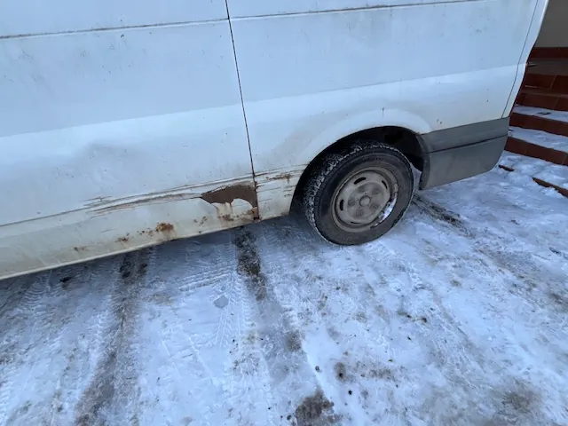 Zbliżenie na dolną część białego vana zaparkowanego na śniegu. Na dolnej krawędzi nadwozia, przed tylnym kołem, widoczne są znaczne uszkodzenia lakieru oraz rdza. Koło jest zabrudzone, a podłoże pokryte jest białym śniegiem z widocznymi śladami opon.