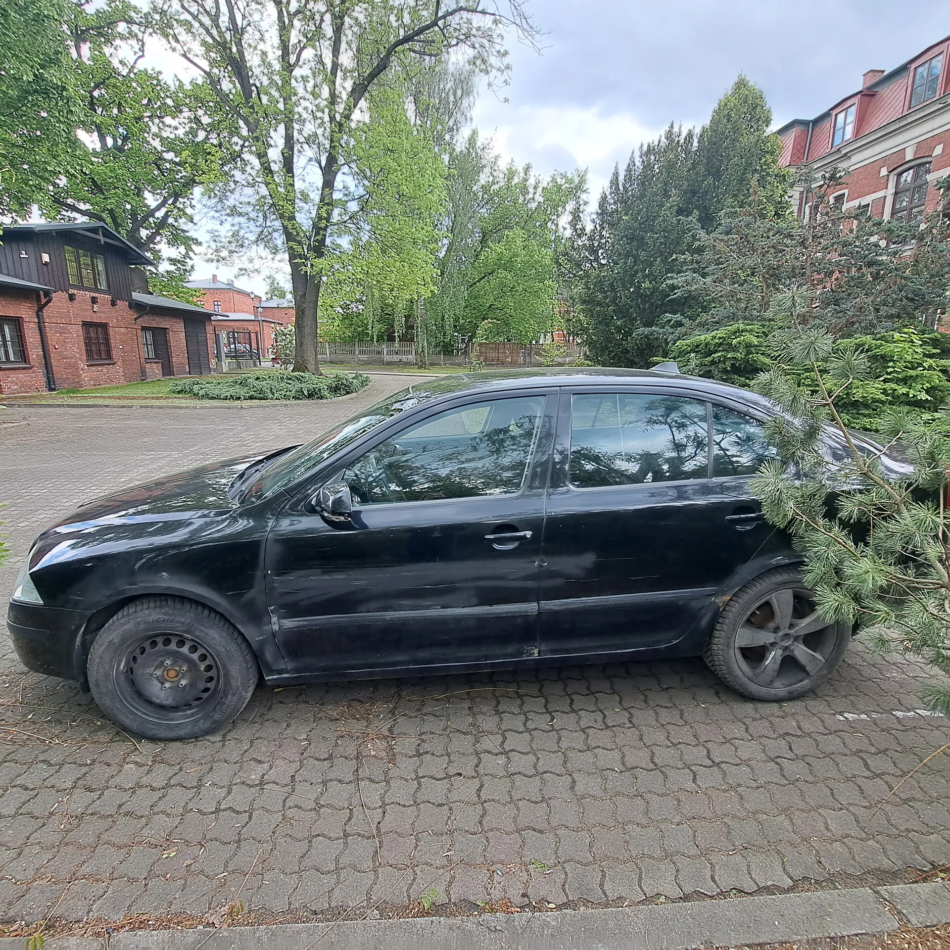 Czarny samochód osobowy typu sedan zaparkowany na brukowanej powierzchni. Po lewej stronie widoczne są ceglane budynki z ciemnymi dachami, po prawej drzewa iglaste i liściaste. W tle drzewa, ogrodzenie oraz ceglany budynek z czerwonym dachem. Niebo częściowo zachmurzone. Samochód ma zużyte opony i widoczne ślady zużycia na karoserii.