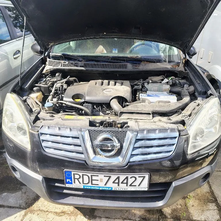 Czarny samochód marki Nissan z otwartą maską, ukazującą silnik i komponenty mechaniczne. Na przodzie pojazdu widoczny jest srebrny grill z logo Nissan oraz tablica rejestracyjna o numerze RDE 74427 z oznaczeniem PL. Poniżej tablicy znajduje się napis: AUTO KOMIS www.mallas-gratka.pl OLSZYNY.