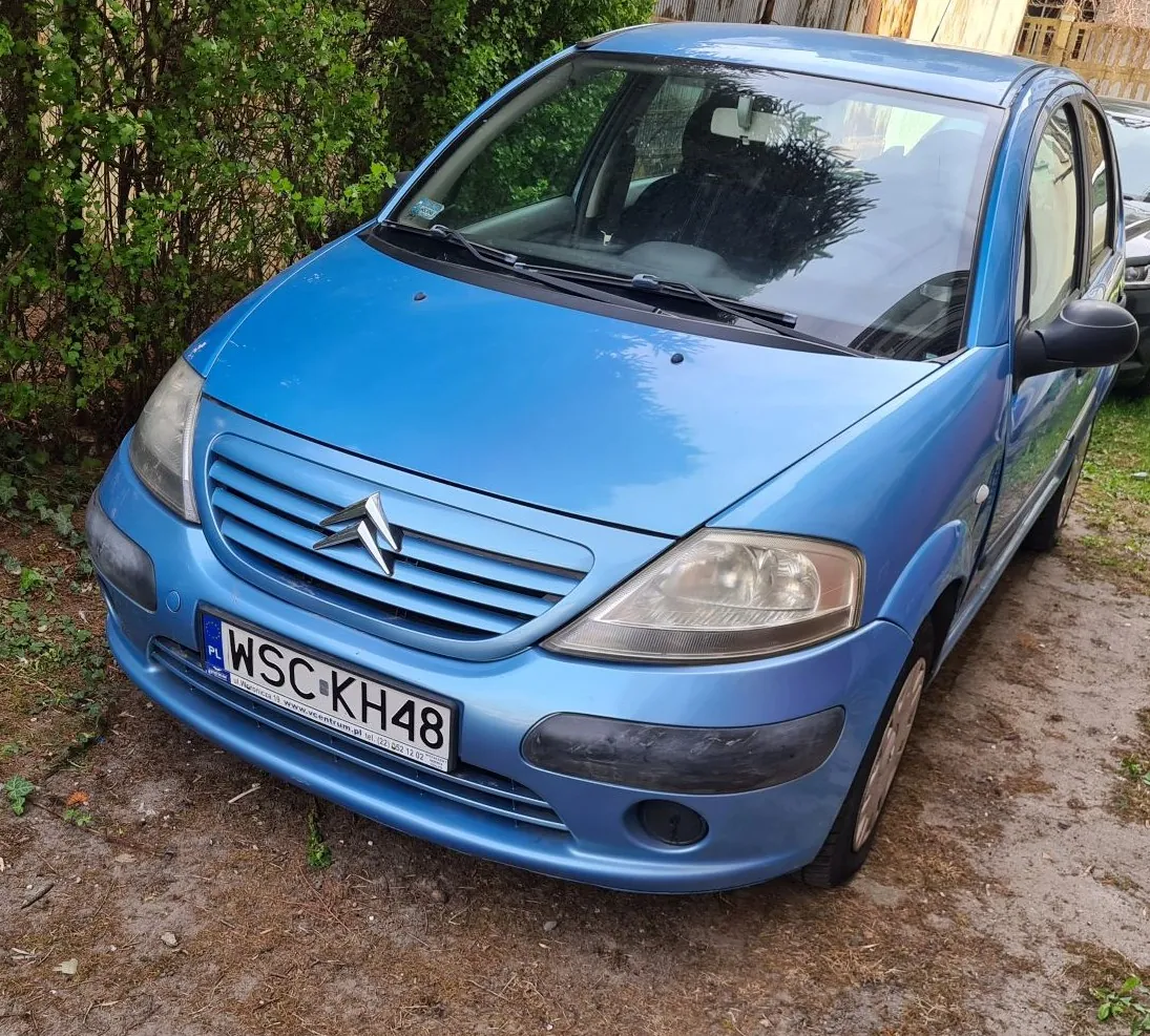 Niebieski samochód marki Citroën zaparkowany na podwórku z ziemią i suchą trawą. Widoczna tablica rejestracyjna z polskim kodem PL oraz numerem WSC KH48. W tle zielone krzewy i drewniany płot. Samochód ma czystą przednią szybę, reflektory i charakterystyczny emblemat marki na masce.