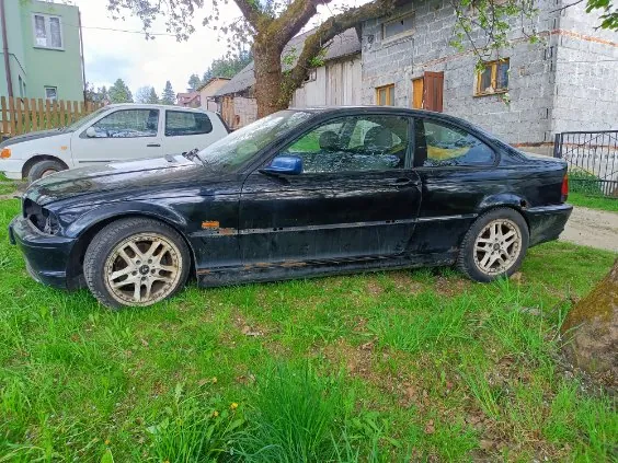 Czarny samochód osobowy typu sedan zaparkowany na trawie. Pojazd ma brudną karoserię z widocznymi uszkodzeniami na boku, złote felgi i nieco przekręconą przednią oponą. W tle widoczny jest biały samochód, zielony dom z drewnianym ogrodzeniem oraz szary budynek z kamienia z drzwiami i oknami. Przed budynkiem rośnie drzewo, a na trawie znajdują się żółte kwiaty. Niebo jest częściowo zachmurzone.