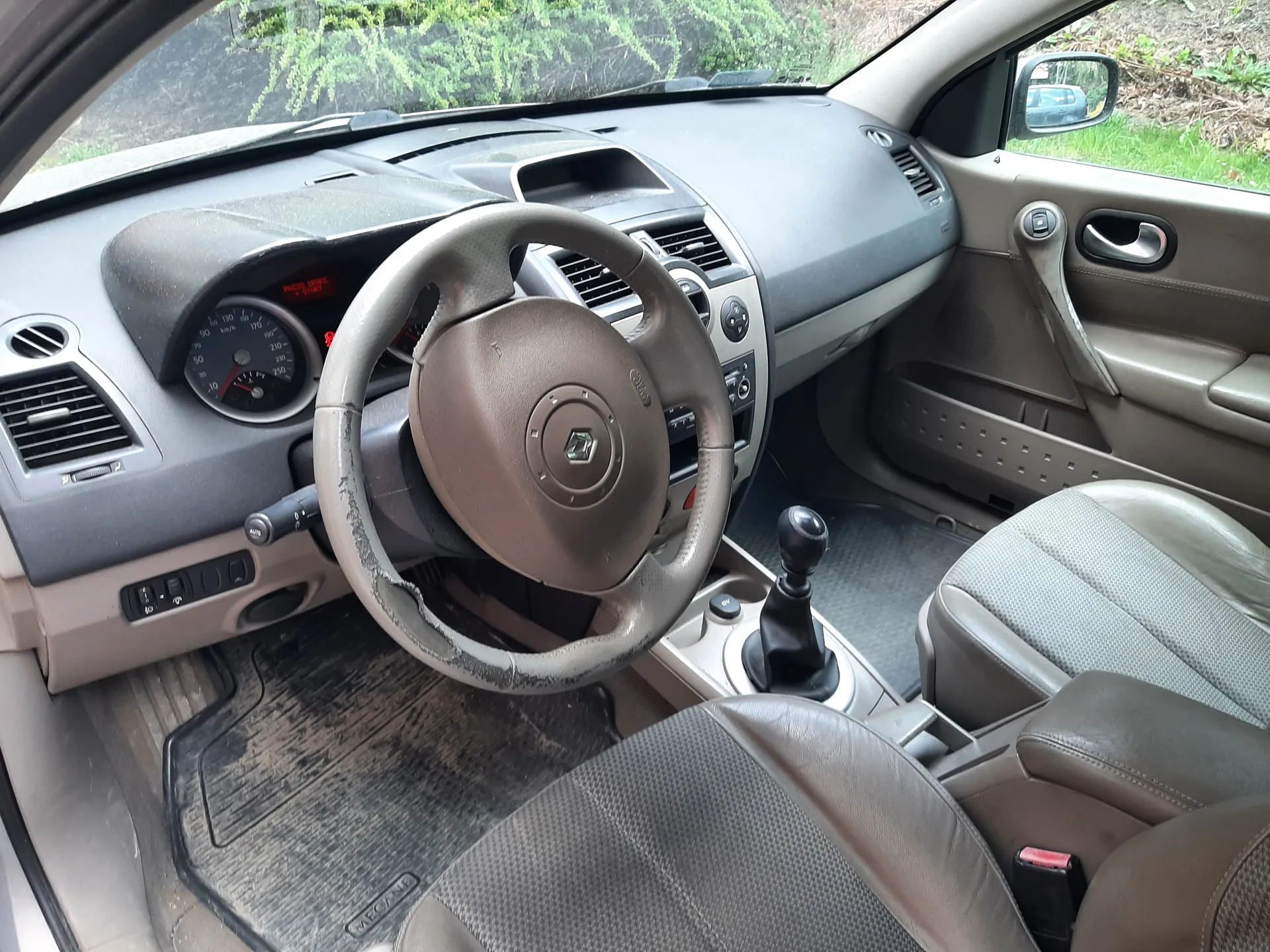 Wnętrze samochodu z widokiem na kierownicę, deskę rozdzielczą i przednie siedzenia. Kierownica w kolorze brązowym z logo w środku, z lewej strony zegar prędkościomierza z czerwonymi wskaźnikami. Deska rozdzielcza w odcieniach szarości i beżu, z widocznymi wentylacjami i przyciskami. Skrzynia biegów z czarną dźwignią umieszczona w środku konsoli. Przednie siedzenia w kolorze szaro-brązowym z tkaniną w drobny wzór. W tle przez przednią szybę widoczne zielone krzewy i fragment innego pojazdu. Drzwi pasażerskie z uchwytem i oknem bocznym.