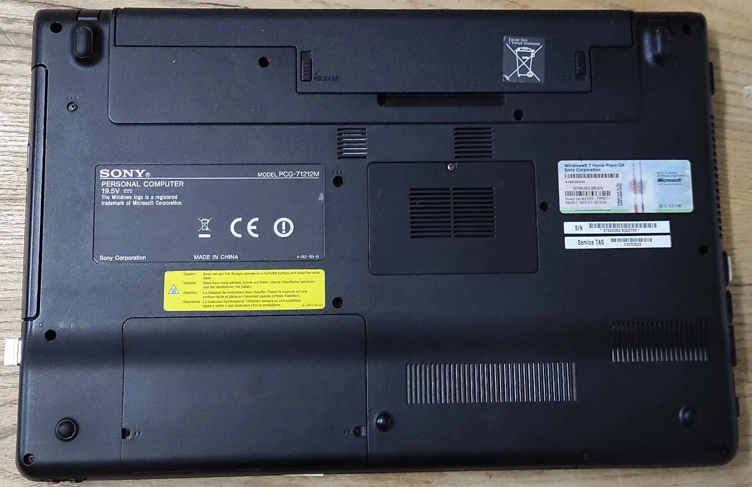 Zdjęcie dolnej obudowy czarnego laptopa marki Sony. Na obudowie widoczne są dwie naklejki. Pierwsza, większa, zawiera logo SONY, napis 'PERSONAL COMPUTER', numer modelu 'MODEL PCG-71212M', oznaczenia CE oraz informację 'MADE IN CHINA'. Druga, mniejsza naklejka zawiera informacje o systemie Windows 7 Home Premium, logo Microsoft oraz kody seryjne i Service TAG. W dolnej części znajduje się żółta naklejka z ostrzeżeniami w kilku językach. Obudowa posiada otwory wentylacyjne oraz gumowe stopki.