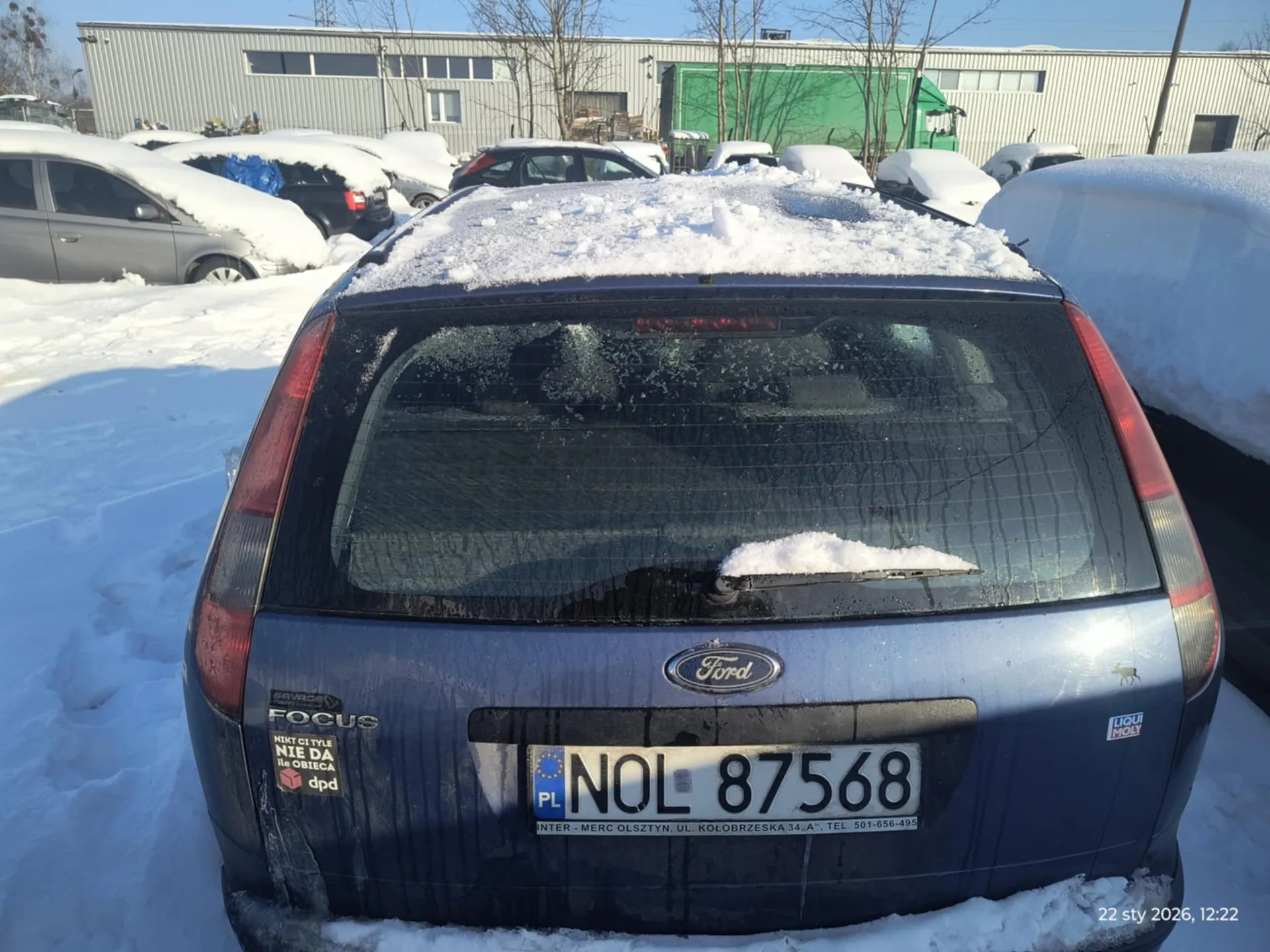 Zdjęcie przedstawia tył niebieskiego samochodu Ford Focus pokrytego śniegiem, zaparkowanego na śnieżnym parkingu. Na tablicy rejestracyjnej widoczny jest numer PL NOL 87568 oraz napisy: "INTER - MERC OLSZTYN, UL. KOŁOBRZESKA 34, A", TEL. 501-656-495". Na bagażniku znajdują się naklejki: "FOCUS", "NIKT CI TYLE NIE DA ile OBIĘCA dpd" oraz logo "LIQUI MOLY". W tle widoczne są inne samochody zakryte śniegiem, przemysłowe budynki, drzewa bez liści i zielony pojazd. W prawym dolnym rogu zaznaczona data i godzina: "22 sty 2026, 12:22". Całość sfotografowana w słoneczny dzień.