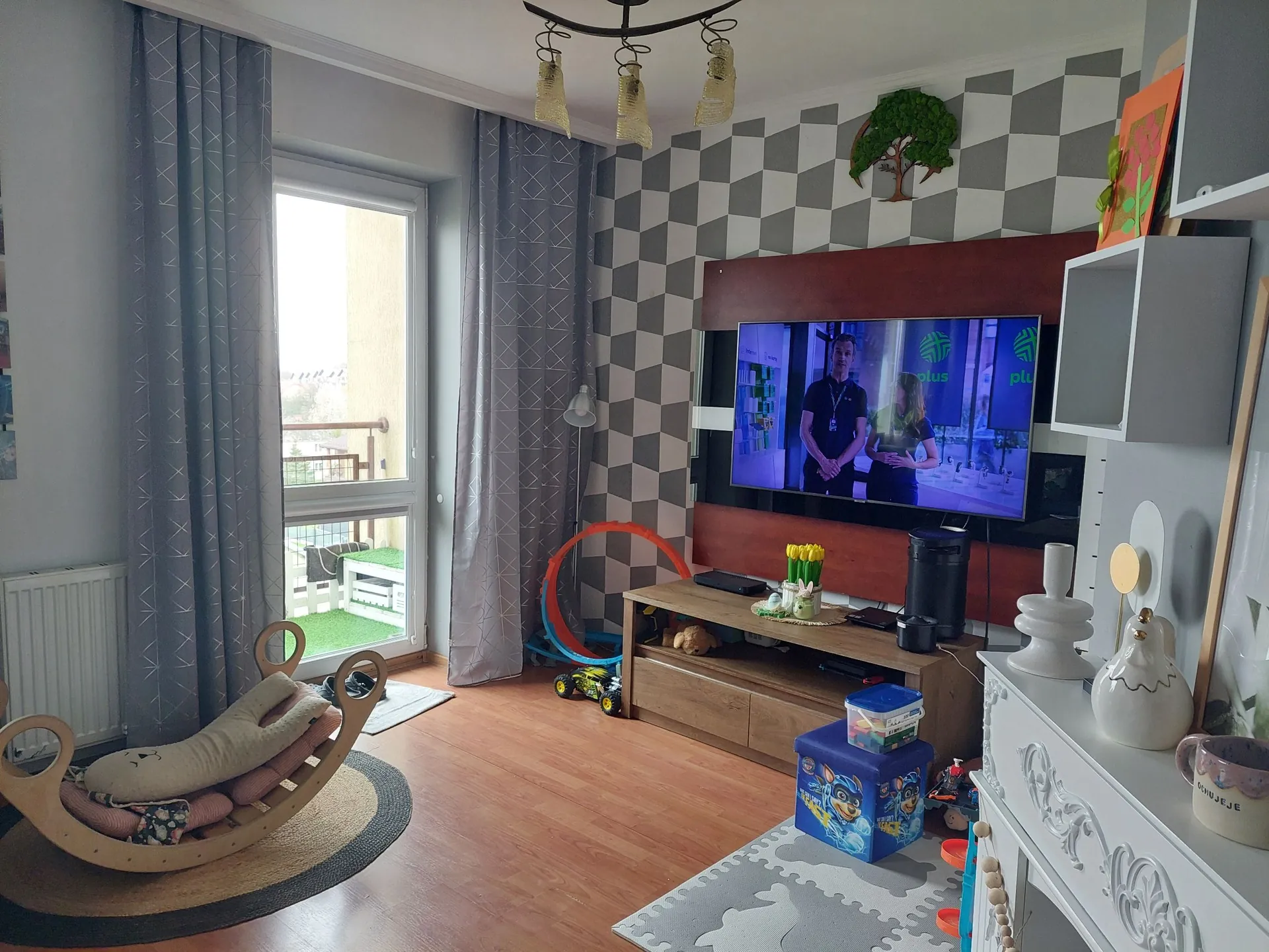 Wnętrze pokoju z drewnianą podłogą. Po lewej stronie znajduje się okno z szarymi zasłonami oraz kołyska dla niemowlęcia na okrągłym dywaniku. Ściana naprzeciwko okna ma wzór szachownicy w odcieniach szarości i bieli. Na ścianie zamontowany jest ciemnobrązowy panel z wbudowanym telewizorem, na którym widoczne są dwie osoby. Pod telewizorem stoi niska drewniana szafka RTV z zabawkami, w tym kolorowy samochód i niebieski organizer. W prawym dolnym rogu widoczny jest fragment białego kominka z dekoracjami, w tym ceramiczna figurka kury i kubek z napisem 'DZIĘKUJĘ'. Z sufitu zwisa lampa z kilkoma kloszami.