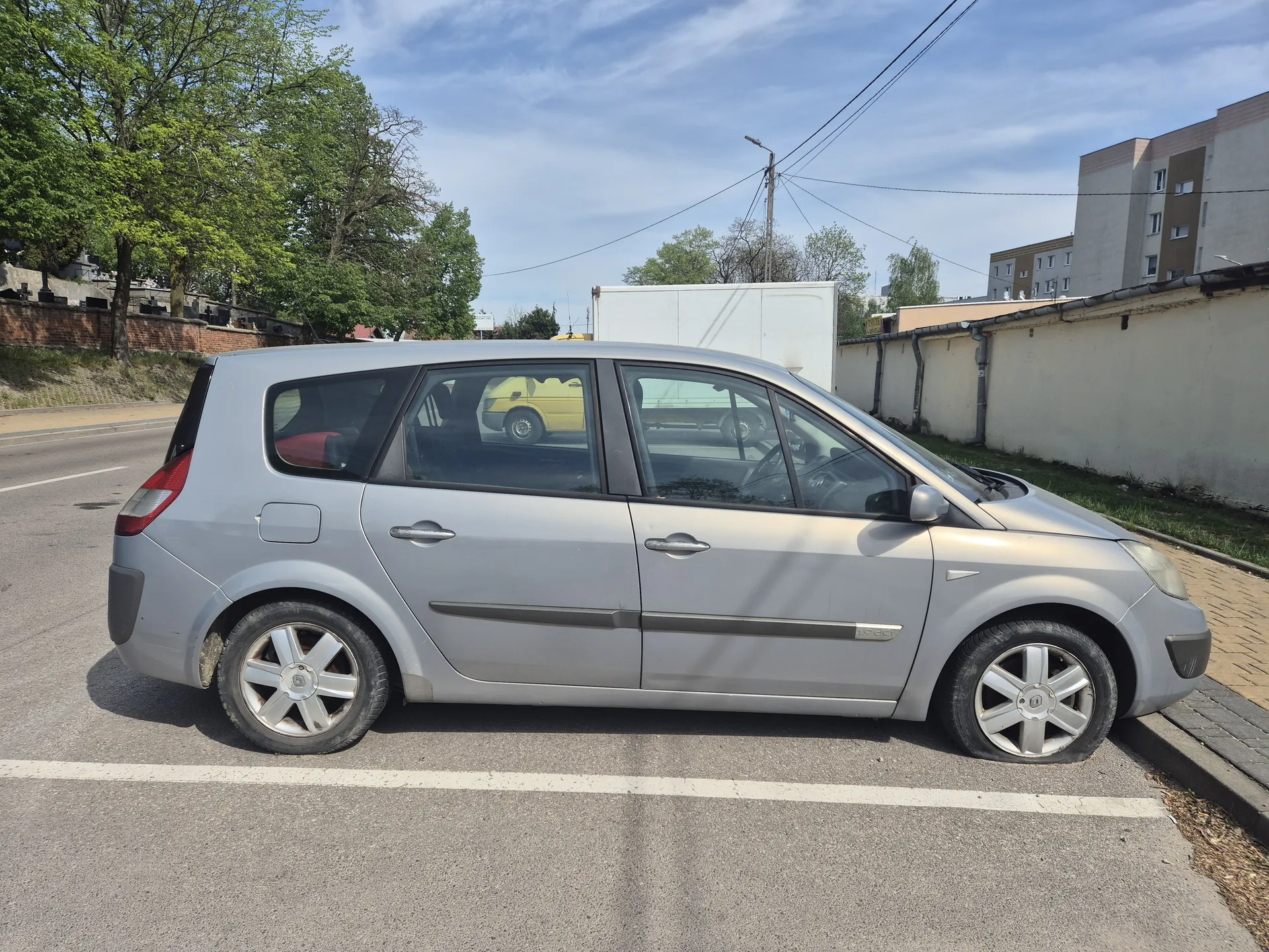 Srebrny samochód osobowy typu minivan zaparkowany na poboczu drogi. Widok z boku, z widocznymi czterema drzwiami, srebrnymi felgami i czarnymi pasami dekoracyjnymi. W tle: drzewa, ceglana ściana z nagrobkami, budynek przemysłowy, żółty samochód, bloki mieszkalne oraz niebo z chmurami. Droga asfaltowa z białą linią wyznaczającą pas ruchu.