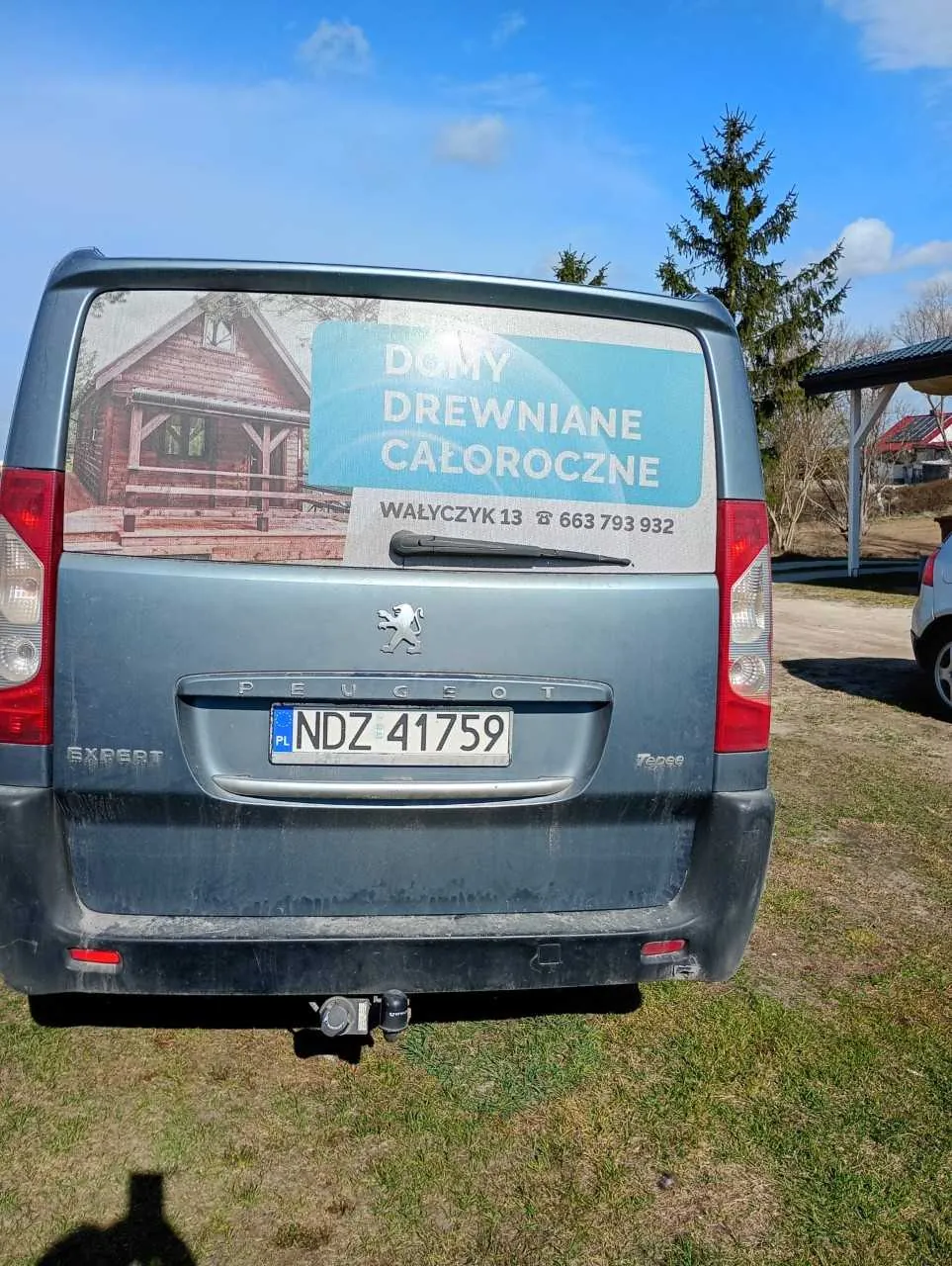 Tył szarego furgonu Peugeot Expert z widocznym logo marki i napisem "EXPERT" po lewej stronie oraz "Tepee" po prawej. Na tylnej szybie naklejka z obrazem drewnianego domu i tekstem: "DOMY DREWNIANE CAŁOROCZNE", "WAŁYCZYK 13" oraz numerem telefonu "663 793 932". Tablica rejestracyjna oznaczona jako "PL NDZ 41759". Furgon stoi na trawie, w tle niebo z chmurami, drzewa i fragment innego pojazdu po prawej stronie.