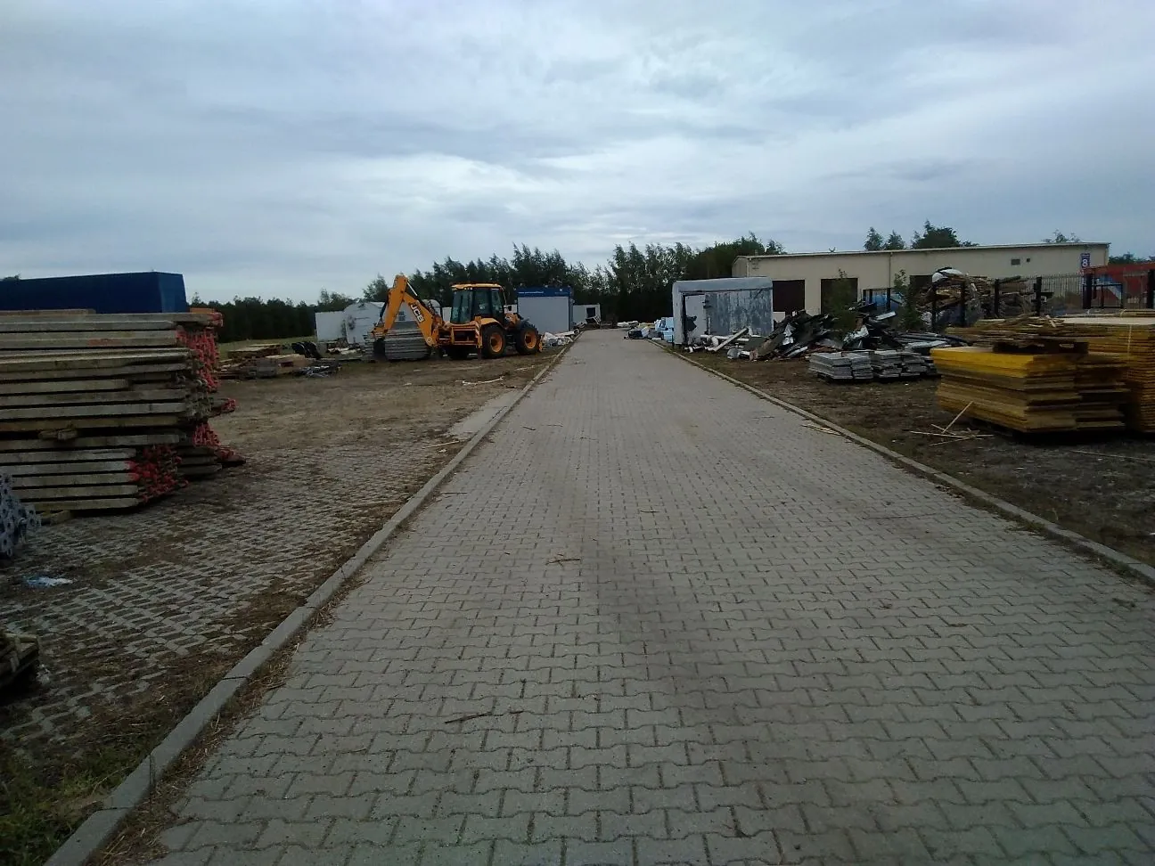 Widok na plac budowy lub składowisko materiałów. W centrum znajduje się szeroka droga z szarej kostki brukowej prowadząca w głąb obrazu. Po lewej stronie widać wysokie stosy drewnianych belek. W oddali stoi żółta koparka ładowacz oraz niebieski kontener. Po prawej stronie znajdują się stosy żółtych płyt oraz inne materiały budowlane, a w tle niski, jasny budynek gospodarczy. Niebo jest zachmurzone.