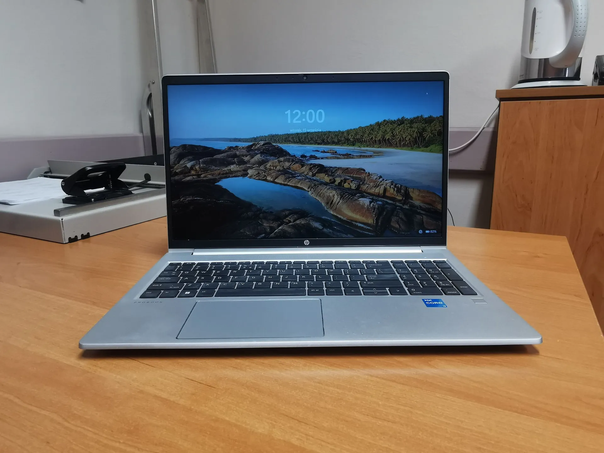 Srebrny laptop marki HP otwarty i ustawiony na drewnianym blacie. Na ekranie widoczna jest tapeta przedstawiająca nadmorski krajobraz z palmami i skałami oraz godzina 12:00. Obok laptopa po lewej stronie znajduje się szara teczka z dokumentami i czarnym zszywaczem. W tle po prawej stronie widoczny jest fragment drewnianej szafki oraz biały czajnik elektryczny.