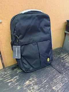 Czarny plecak z przodem kieszeni i małym żółtym logo, umieszczony na zużytej drewnianej powierzchni. Obok widoczna brązowa powierzchnia kartonowa i fragment zielonego przedmiotu w lewym górnym rogu.