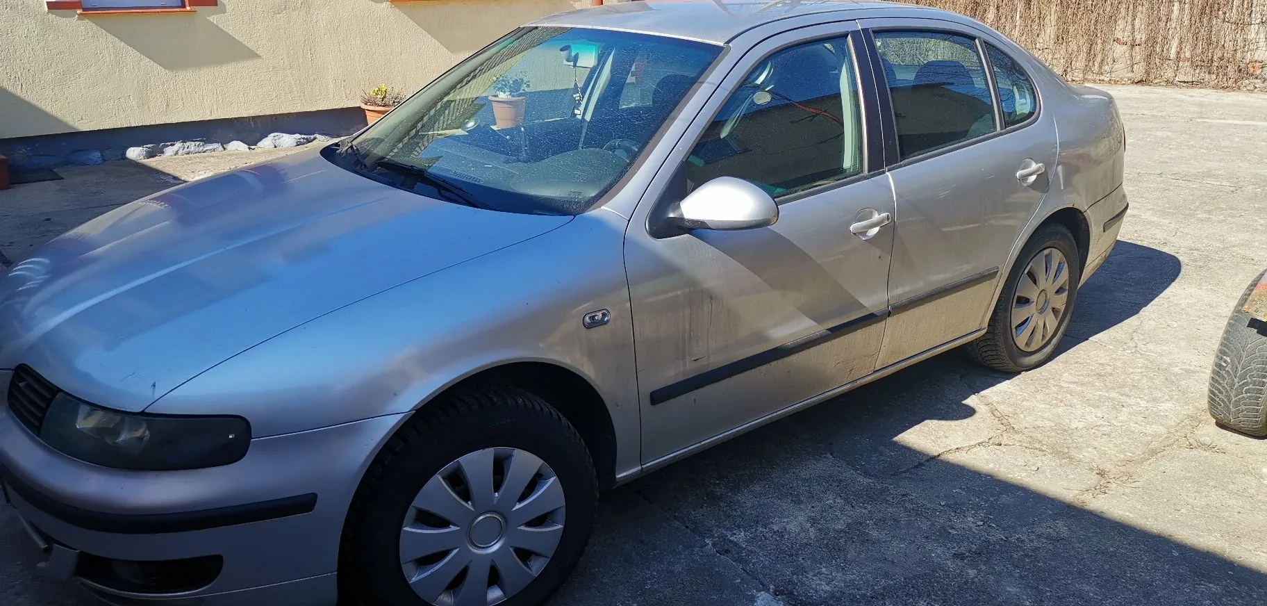 Srebrny samochód osobowy typu sedan zaparkowany na betonowym podjeździe przed jasnym budynkiem. Pojazd jest widoczny z profilu, z przodu i z boku, w pełnym słońcu.