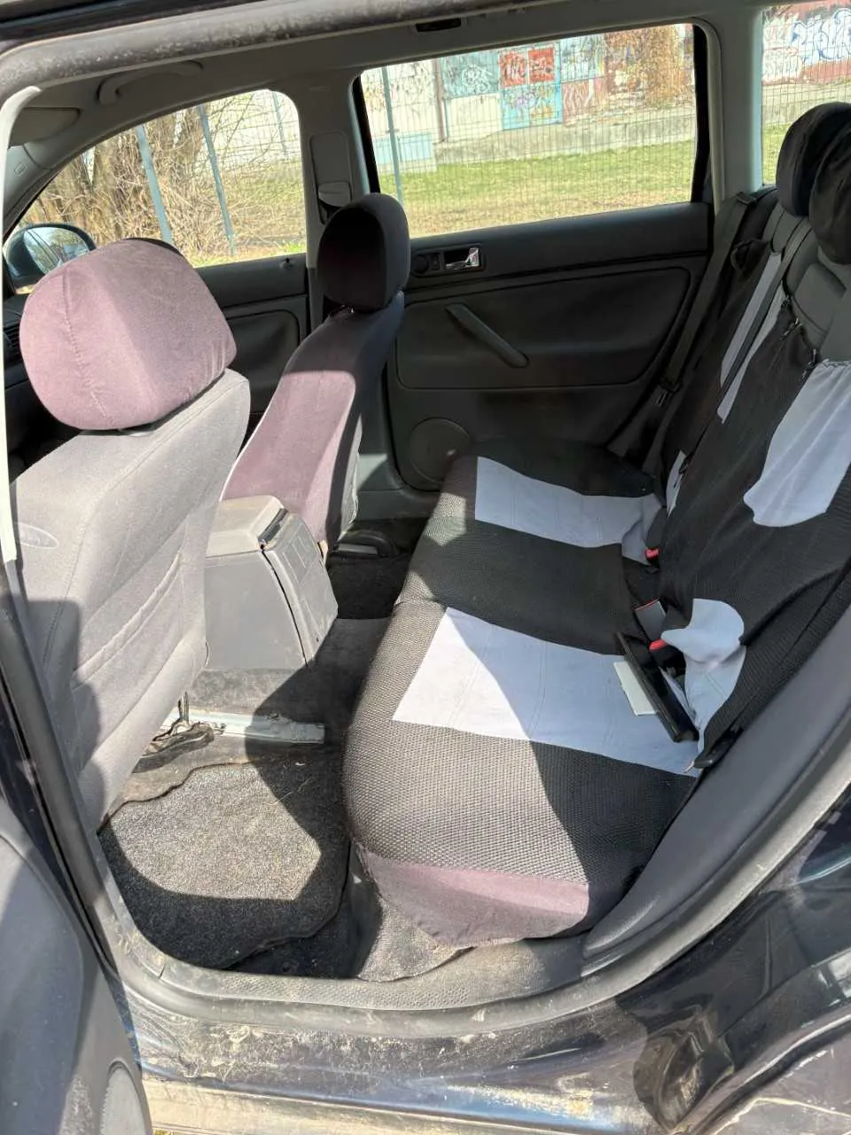 Wnętrze samochodu widziane od strony otwartego tylnego drzwi. Widoczne są dwa tylne siedzenia z pokrowcami w kolorze szarym i fioletowym, poduszki zagłówkowe oraz pasy bezpieczeństwa. Na podłodze znajduje się uszkodzony dywanik z widoczną dziurą. Poza samochodem przez okna widać siatkę ogrodzeniową, trawę oraz budynek z kolorowymi graffiti. Wnętrze drzwi jest czarne, z widocznym uchwytem i okienkiem.