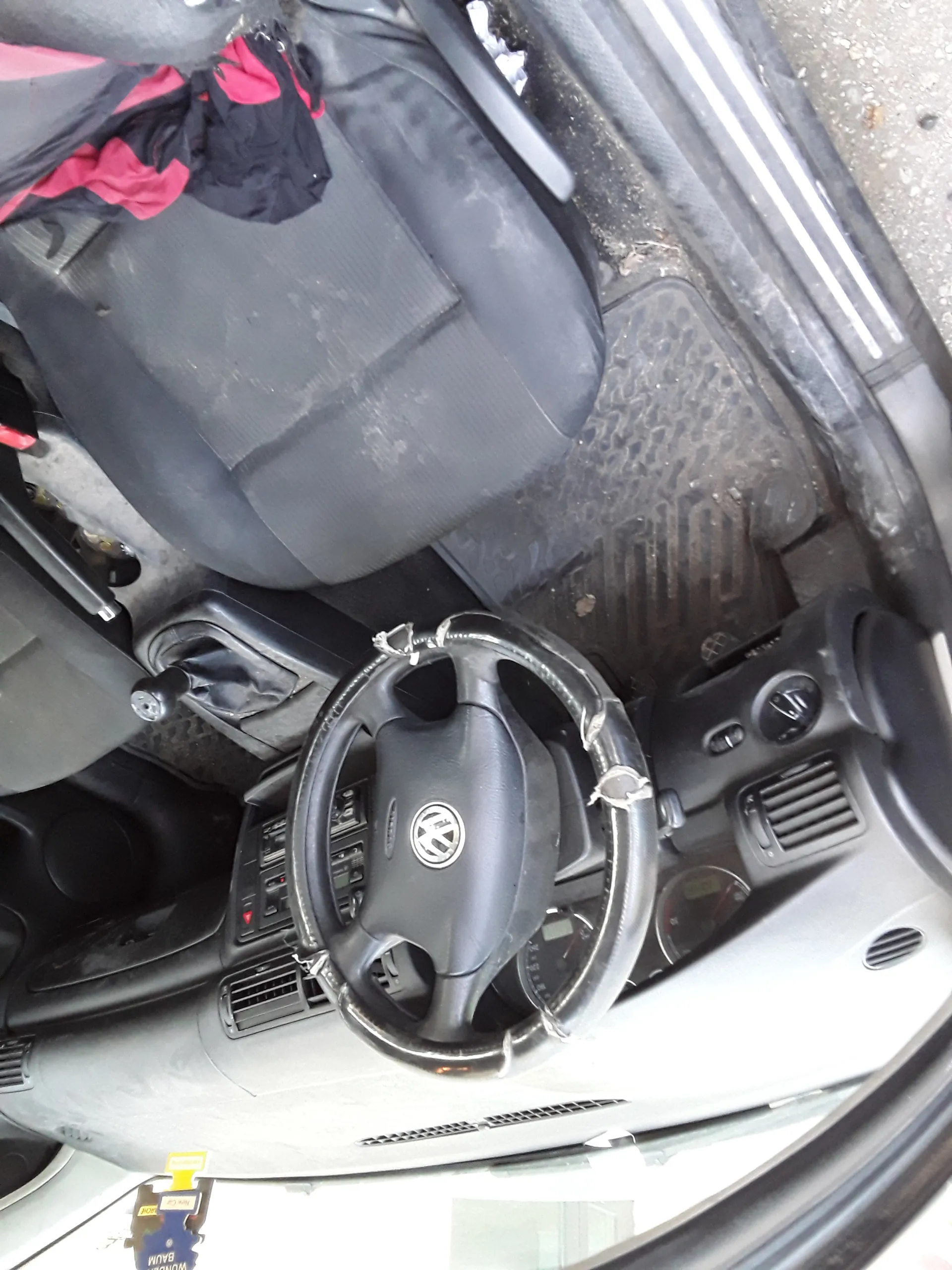 Wnętrze samochodu marki Volkswagen z widoczną kierownicą z logo VW, czarnymi siedzeniami tkaninowymi, brudnymi dywanikami podłogowymi, dźwignią zmiany biegów oraz deską rozdzielczą z wentylacjami i przyciskami. Na siedzeniu pasażera leży czarno-czerwona kurtka. Na przedniej szybie widoczny bilet parkingowy z napisem "PARKING" i "EXPIRED".