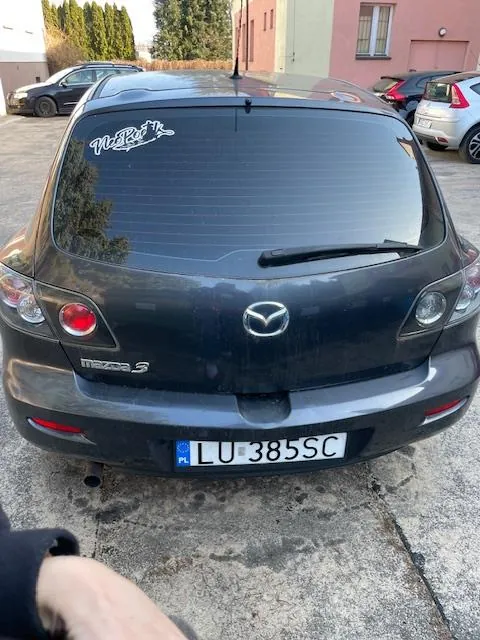 Z tyłu czarny samochód Mazda 3 z tablicą rejestracyjną LU-385SC (PL) umieszczoną na środku. Na szybie tylnej naklejka z napisem "NARL" w stylizowanym czcionku. W tle widoczne inne zaparkowane pojazdy, różowe i beżowe budynki oraz drzewa. Na dole zdjęcia fragment ręki w czarnej rękawicy. Samochód stoi na betonowym placu parkingowym.