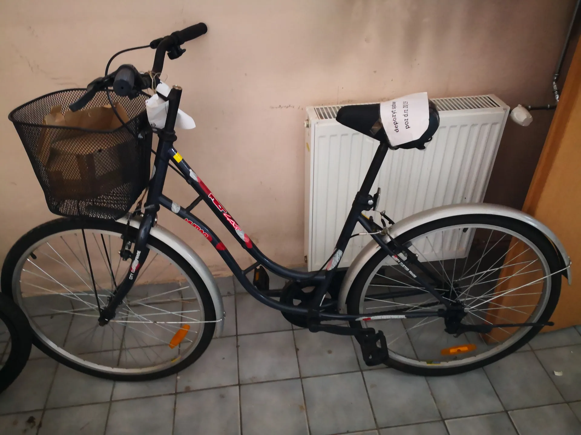 Ciemnoniebieski rower z czerwonymi akcentami, z czarnym siatkowym koszem na przednim kole zawierającym brązową torbę papierową. Na siodełku biały znaczek z napisem '500 zł 2019 depozyt'. Rower oparty o beżową ścianę obok białego grzejnika, na szarych płytkach podłogowych. Widoczne czarne uchwyty kierownicy i pomarańczowe odblaski na kołach.