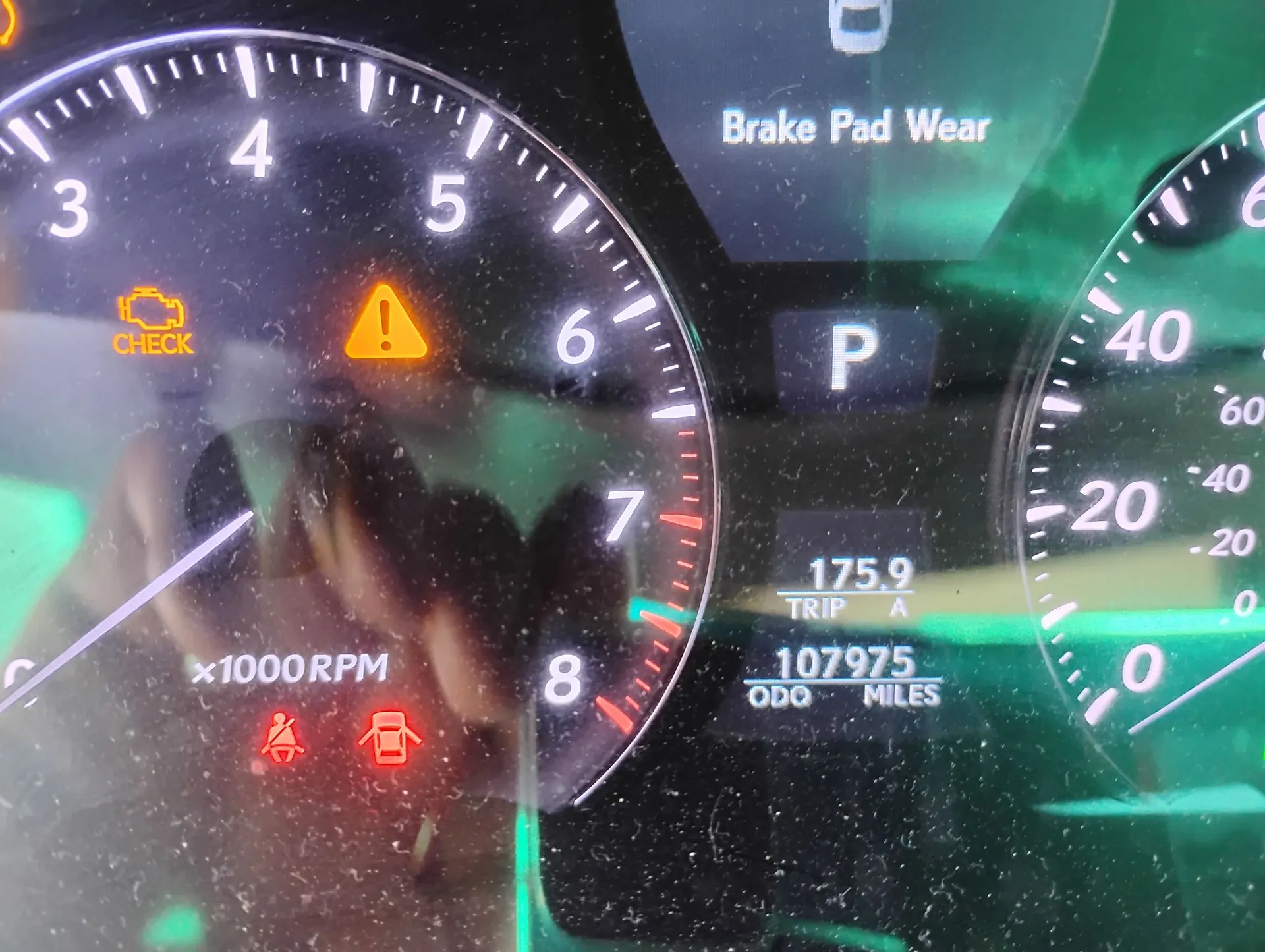 Zbliżenie na deskę rozdzielczą samochodu. Widoczny jest obrotomierz z cyframi od 3 do 8 i napisem 'x1000RPM'. Na wyświetlaczu zapalone są pomarańczowe kontrolki: ikona silnika z napisem 'CHECK' oraz trójkąt z wykrzyknikiem. Na dole widoczne są czerwone ikony niezapiętych pasów bezpieczeństwa oraz otwartych drzwi. Po prawej stronie znajduje się wskaźnik biegu 'P', licznik przebiegu 'ODO 107975 MILES' oraz licznik podróży 'TRIP A 175.9'. W górnej części ekranu widnieje napis 'Brake Pad Wear'. Po prawej stronie widoczny jest fragment prędkościomierza.