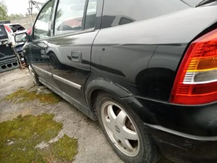 Czarny samochód osobowy widziany z boku, z tyłu. Widoczne są czerwono-żółte światło tylnego reflektora, srebrna felga z logo oraz fragment asfaltowego podłoża z plamami mchu. W tle częściowo widoczny inny pojazd z otwartą maską oraz budynki.