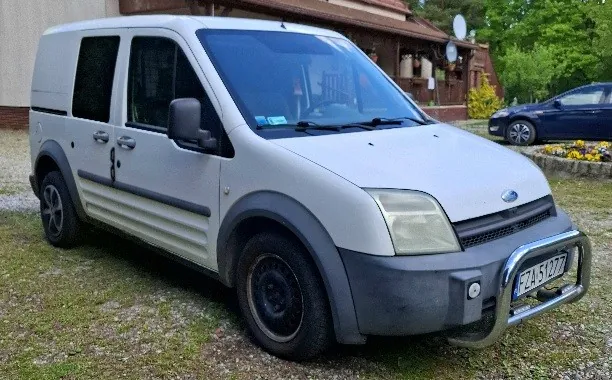 Biały samochód dostawczy marki Ford z szarym dolnym elementem nadwozia, zaparkowany na podwórku z żwirem i trawą. Widoczna tablica rejestracyjna FZ51977 oraz metalowy zderzak przedni. W tle dom z brązowym dachem, satelity, ciemnoniebieski samochód osobowy oraz zieleń z drzewami i kwiatami.