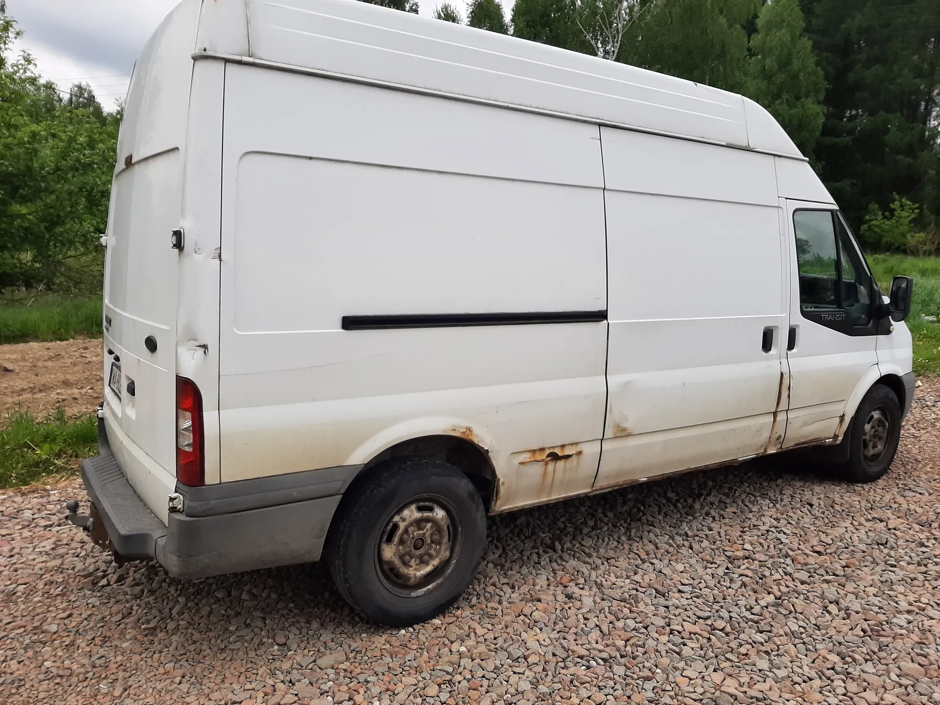 Biały dostawczy van Ford Transit z widocznymi śladami rdzy na bokach i dolnej części nadwozia, zaparkowany na żwirowej powierzchni. Van posiada czerwone światło tylne, czarną listwę na boku oraz zużyte opony. W tle zielone drzewa i trawa. Na przedniej szybie widoczny napis 'TRANSIT'.