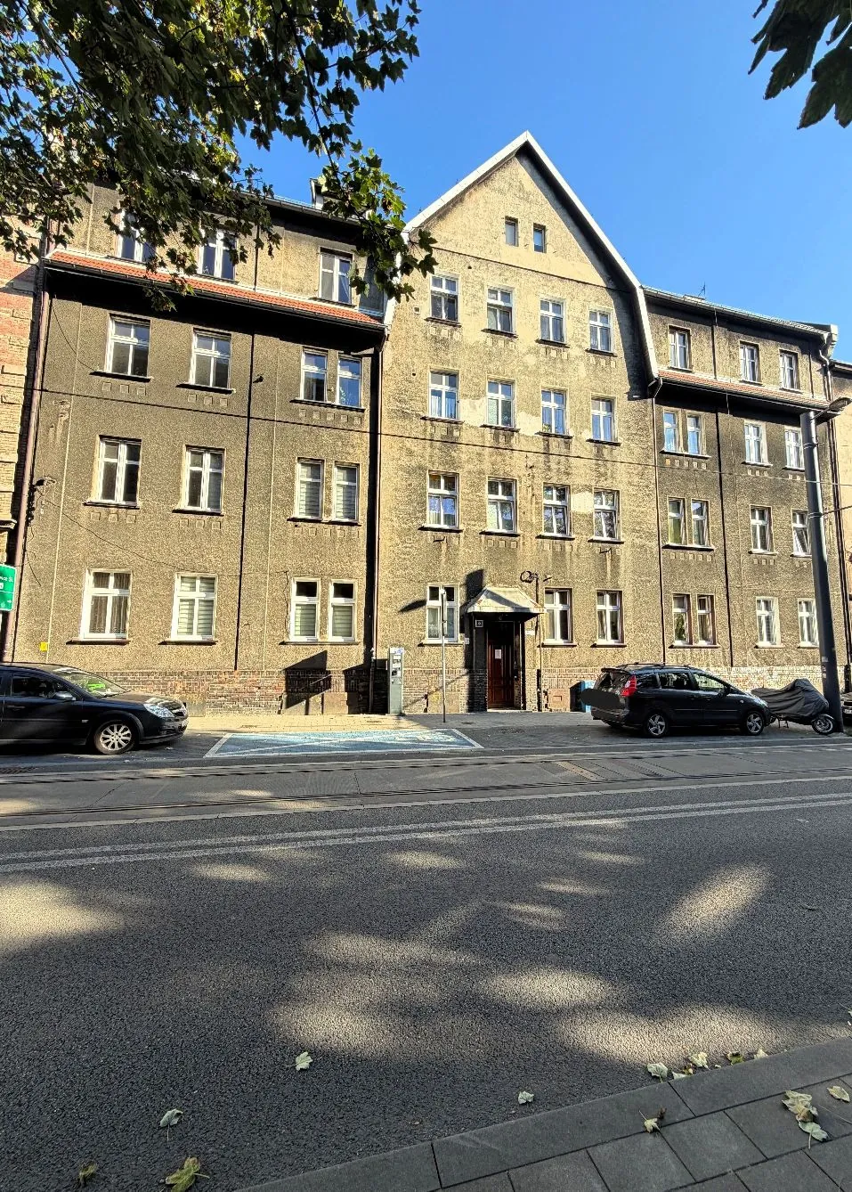 Wielopiętrowy, szary budynek mieszkalny z tynkiem o chropowatej strukturze i spadzistym dachem. Budynek posiada liczne prostokątne okna rozmieszczone w rzędach. Na parterze znajduje się wejście z ciemnymi drzwiami i małym daszkiem. Przed budynkiem przebiega asfaltowa droga z białymi liniami oznakowania poziomego. Po lewej stronie zaparkowany jest ciemny samochód, a po prawej czarny samochód typu kombi. W górnej części zdjęcia widoczne są gałęzie drzew z zielonymi liśćmi na tle bezchmurnego, niebieskiego nieba.