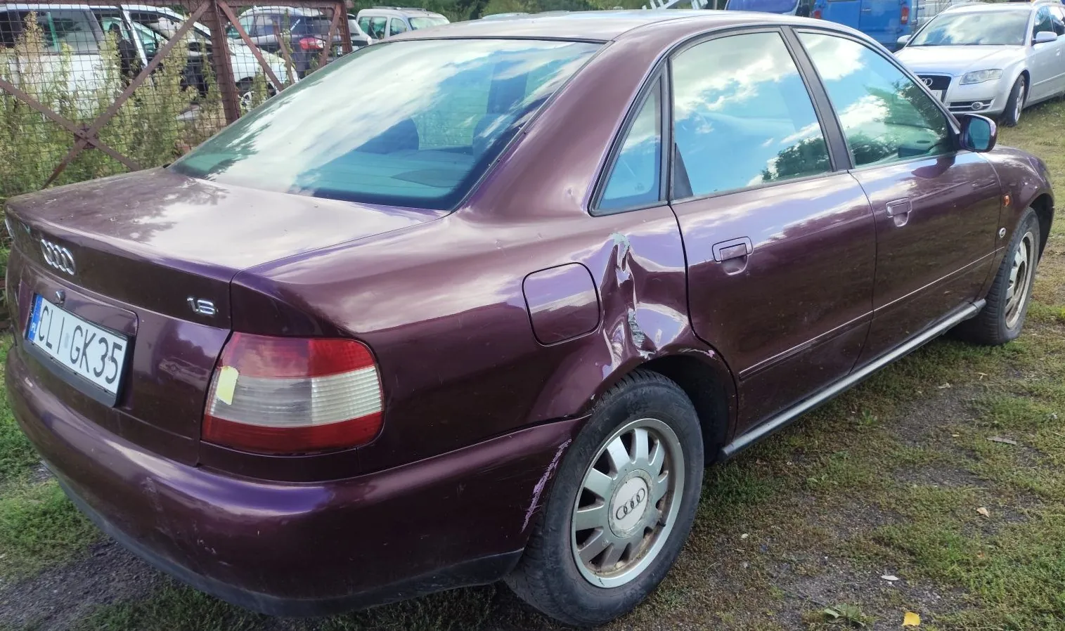 Bordowy samochód marki Audi w widoku z tyłu i z boku. Pojazd posiada widoczne uszkodzenia w postaci wgnieceń na tylnym błotniku nad prawym kołem oraz na tylnym zderzaku. Na klapie bagażnika znajduje się logo Audi, oznaczenie modelu '1.8' oraz tablica rejestracyjna o numerze 'CLI GK 35'. Samochód stoi na trawiasto-żwirowym podłożu, w tle widoczne są inne zaparkowane auta i ogrodzenie z siatki.