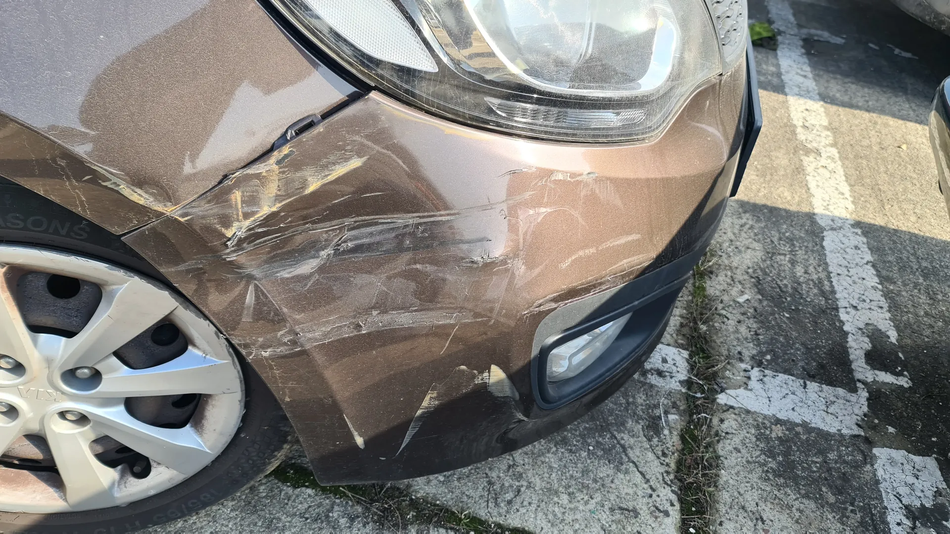 Zbliżenie na przód brązowego samochodu z widocznymi pionowymi i poziomymi zadrapaniami na masce i zderzaku. Widoczne są reflektor, część koła z srebrną felgą oraz fragment opony z częściowo czytelnym napisem SONS. Samochód stoi na betonowej powierzchni z białymi liniami parkingu.