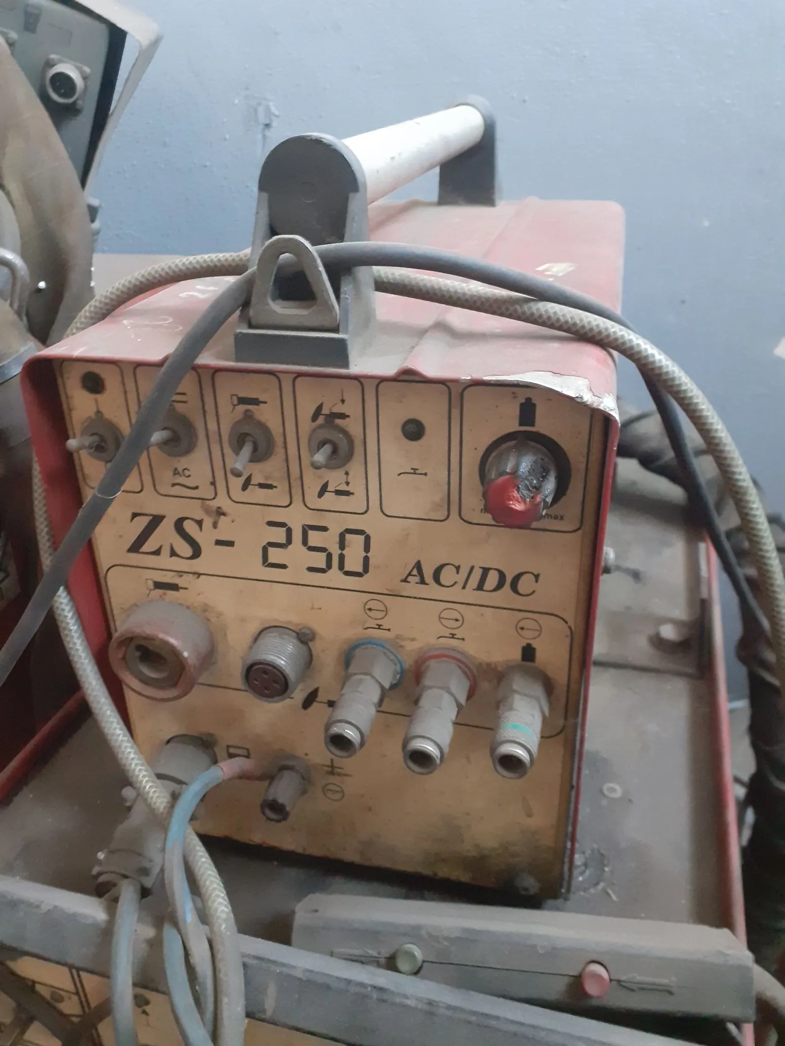 Zdjęcie starej, czerwonej spawarki elektrycznej z oznaczeniem 'ZS- 250 AC/DC'. Urządzenie posiada panel sterowania z kilkoma pokrętłami, przełącznikami oraz gniazdami do podłączenia przewodów. Na panelu widoczne są symbole prądu przemiennego (AC) i stałego (DC). Do urządzenia podłączone są grube, szare kable. Sprzęt jest zakurzony i nosi ślady użytkowania.