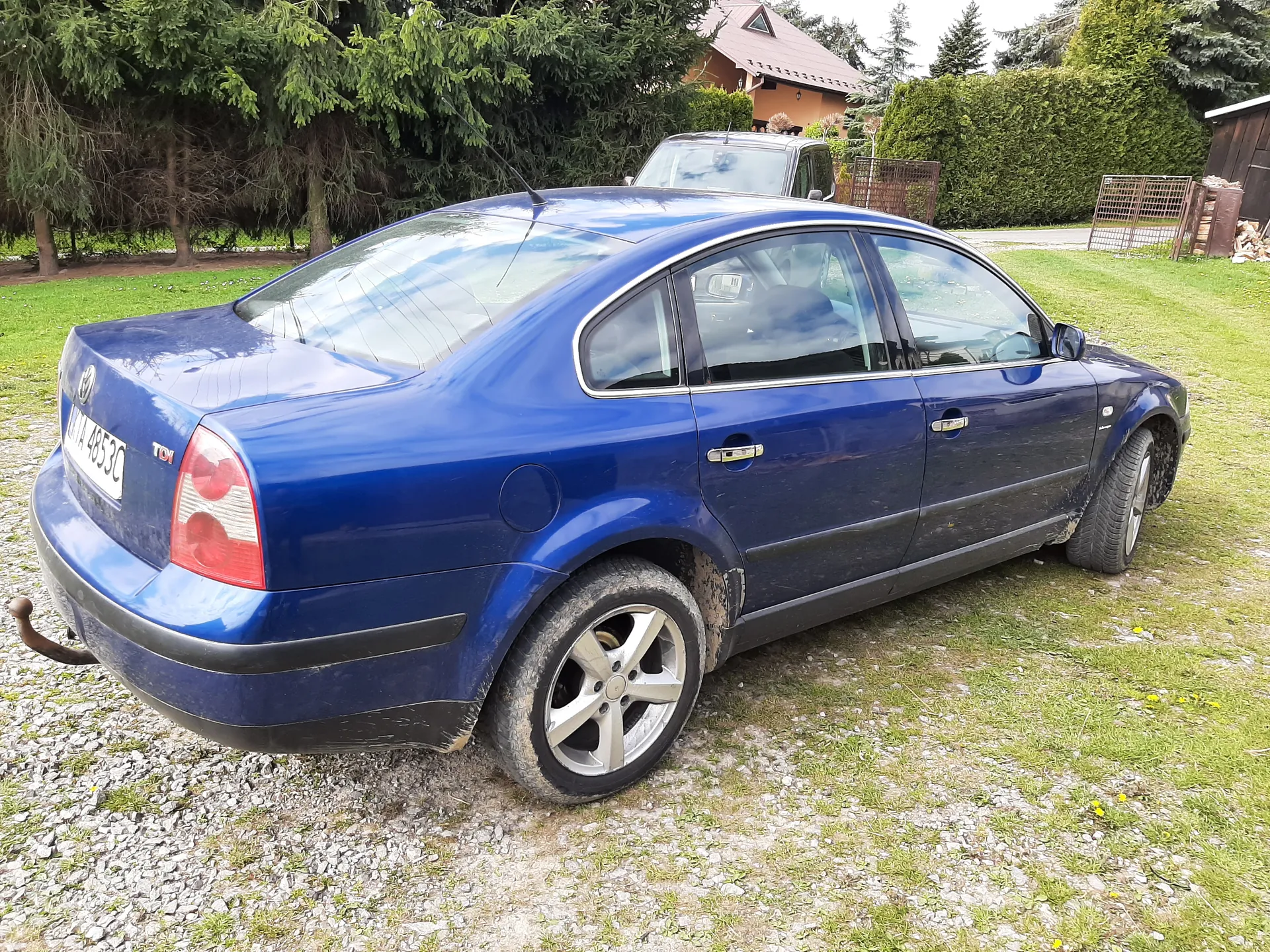 Niebieski samochód osobowy typu sedan zaparkowany na podwórku z żwirem i trawą. Widoczna tablica rejestracyjna z numerem 1A 4853C. Samochód ma zabrudzone koła i dolne części nadwozia. W tle dom z brązowym dachem, zielone krzewy, drzewa oraz drugi pojazd. Po prawej stronie widoczna drewniana szopa z drewnem i metalowe ogrodzenie.