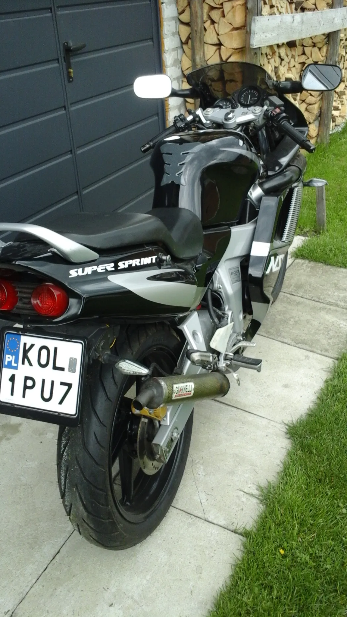 Czarno-srebrny motocykl sportowy leżący na boku na betonowej nawierzchni. Na boku widoczny napis SUPER SPRINT. Tablica rejestracyjna z napisem KOL 1PU7 i oznaczeniem PL. W tle stos drewna, czarna brama garażowa oraz zielona trawa.