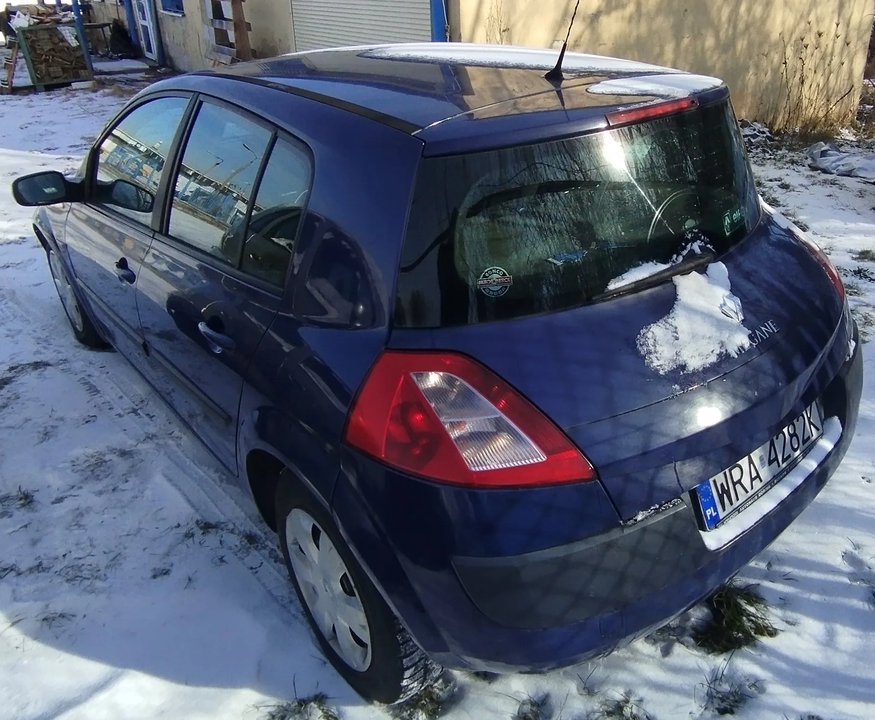 Ciemnoniebieski samochód osobowy typu hatchback zaparkowany na śnieżnej powierzchni. Widok z tyłu i boku. Na tylnym szybie widoczny napis MEGANE oraz naklejka. Tablica rejestracyjna PL WRA 4282K. Na dachu i tylnej części samochodu leży śnieg. W tle budynek z zamkniętą bramą garażową, fragmenty muru i stos drewna. Śnieg pokrywa ziemię, widoczne są ślady opon.
