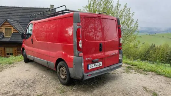 Czerwony furgon marki Opel z czarnym bagażnikiem dachowym, zaparkowany na piaszczystym podjeździe. Pojazd ma polską tablicę rejestracyjną o numerze KR 9NA34. W tle widoczny jest drewniany dom z ciemnym dachem oraz zielone, pagórkowate tereny z drzewami pod zachmurzonym niebem.