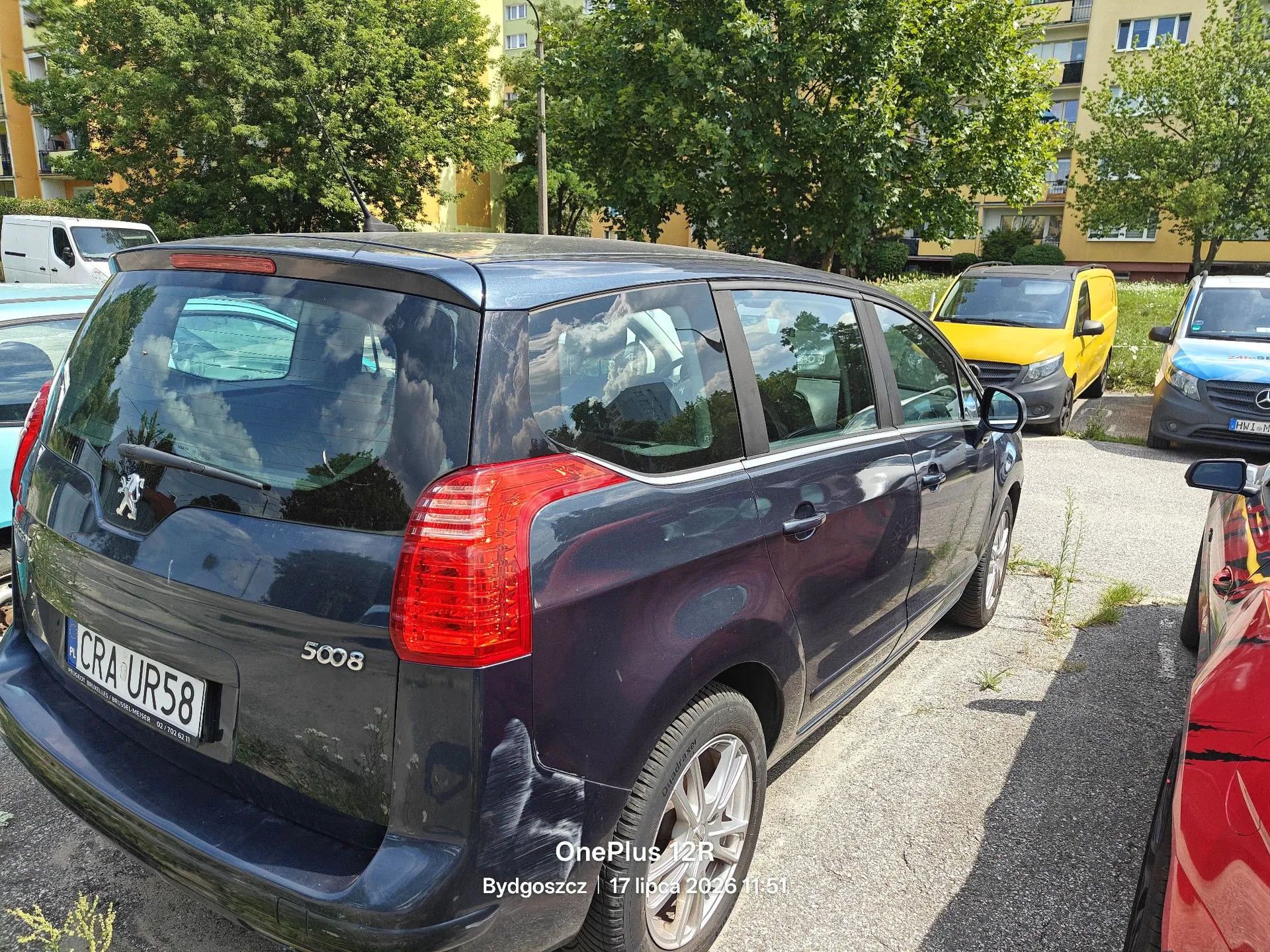 Ciemnoniebieski samochód marki Peugeot 5008 zaparkowany na asfaltowym parkingu. Widoczna jest tylna część i prawy bok auta. Samochód ma tablicę rejestracyjną o numerze CRA UR58. W tle widać zielone drzewa, żółtego vana oraz fragmenty innych samochodów. Na dole obrazu znajduje się biały napis: 'OnePlus 12R Bydgoszcz | 17 lipca 2024 11:51'.