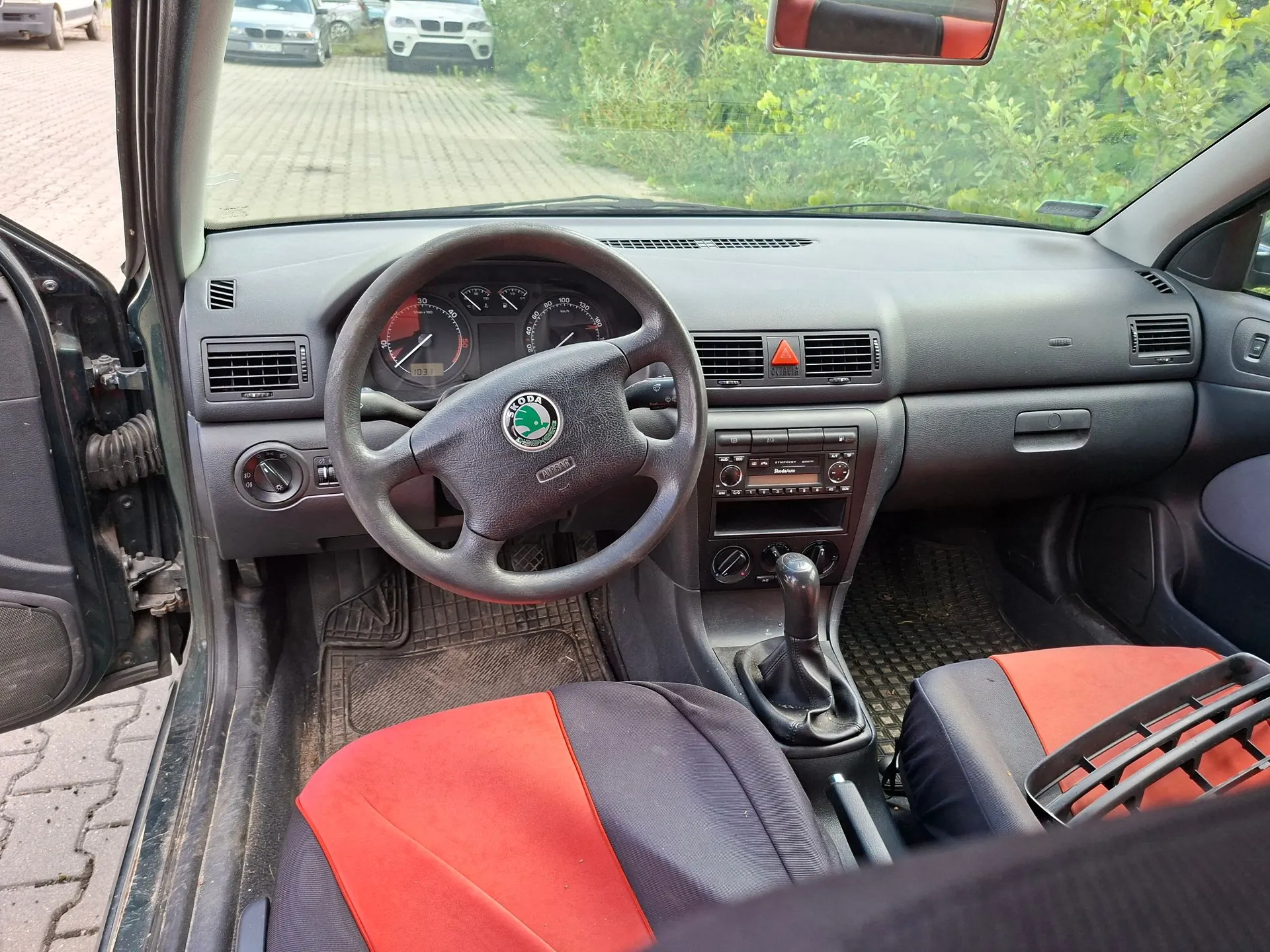 Wnętrze samochodu marki Skoda widziane z perspektywy pasażera. Widoczna jest ciemnoszara deska rozdzielcza, kierownica z logo Skody, zegary analogowe, radio oraz lewarek zmiany biegów. Fotele mają czarno-czerwoną tapicerkę. Przez przednią szybę widać zaparkowane samochody i zieleń.