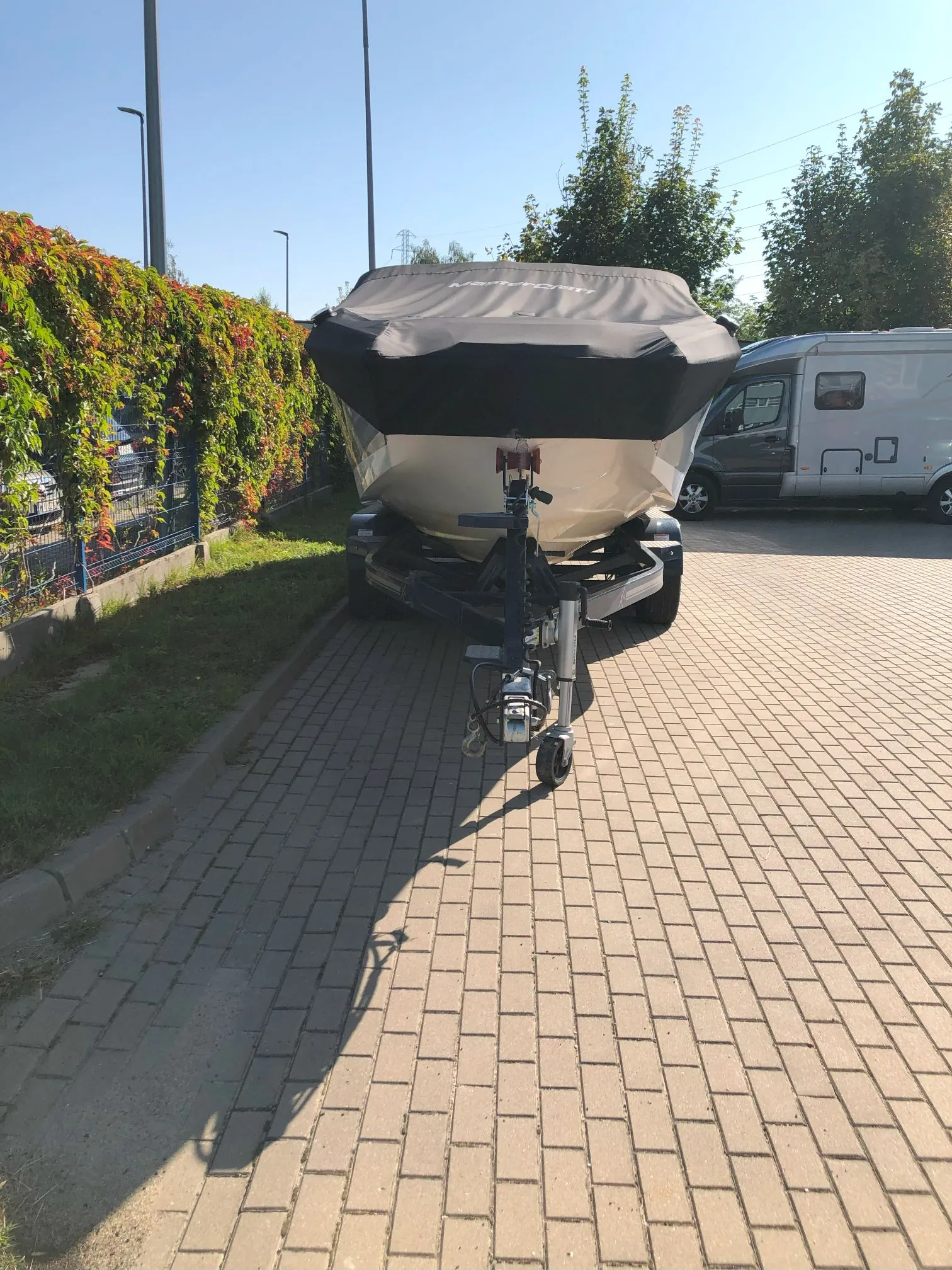 Łódź motorowa przykryta czarną plandeką, ustawiona na przyczepie transportowej. Przyczepa stoi na kostce brukowej obok trawnika i zielonego ogrodzenia z pnączami. W tle widoczny jest srebrny kamper oraz drzewa pod bezchmurnym, niebieskim niebem.
