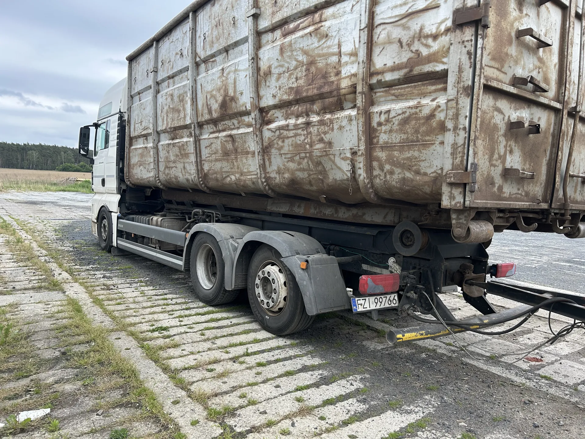 Biały ciężarówka z przyczepą o metalowej, zardzewiałej powierzchni. Widoczna tablica rejestracyjna FZI 99766. Pojazd stoi na nawierzchni z betonowych płyt z trawą między nimi. W tle pole, las i zachmurzone niebo.