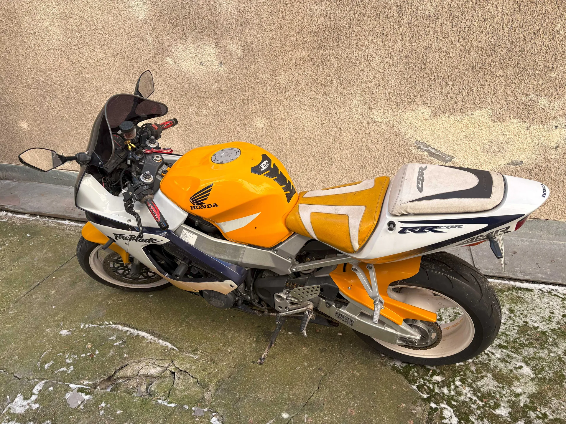 Motocykl marki Honda model FireBlade w kolorze pomarańczowo-białym z niebieskimi akcentami, zaparkowany na betonowej powierzchni z resztkami śniegu. Na zbiorniku paliwa widoczny czarny napis HONDA oraz logo marki, na boku motocykla napis FireBlade, a na części tylnej napis RR. Tło stanowi beżowa, teksturowana ściana. Motocykl posiada czarne koła, lusterka boczne oraz czarny wiatrochron.