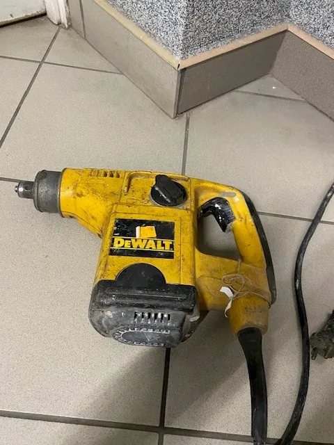 żółta wiertarka udarowa marki DeWALT z widocznymi śladami użytkowania, leżąca na podłodze z kafelkami. W tle schody z granitowym stopniem. Czarny kabel wychodzący z narzędzia. Czytelny napis 'DeWALT' na boku wiertarki.