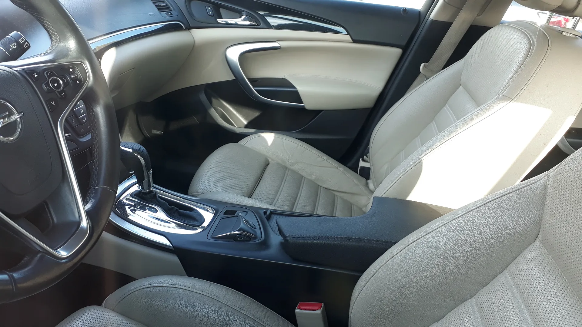 Wnętrze samochodu z widocznym kierownicą z logo, jasnobrązowymi skórzany siedzeniami, centralną konsolą z dźwignią zmiany biegów, czarno-beżowym panelem drzwiowym z uchwytem oraz przyciskami sterującymi na kierownicy. W tle widoczne są fragmenty deski rozdzielczej i pasów bezpieczeństwa.