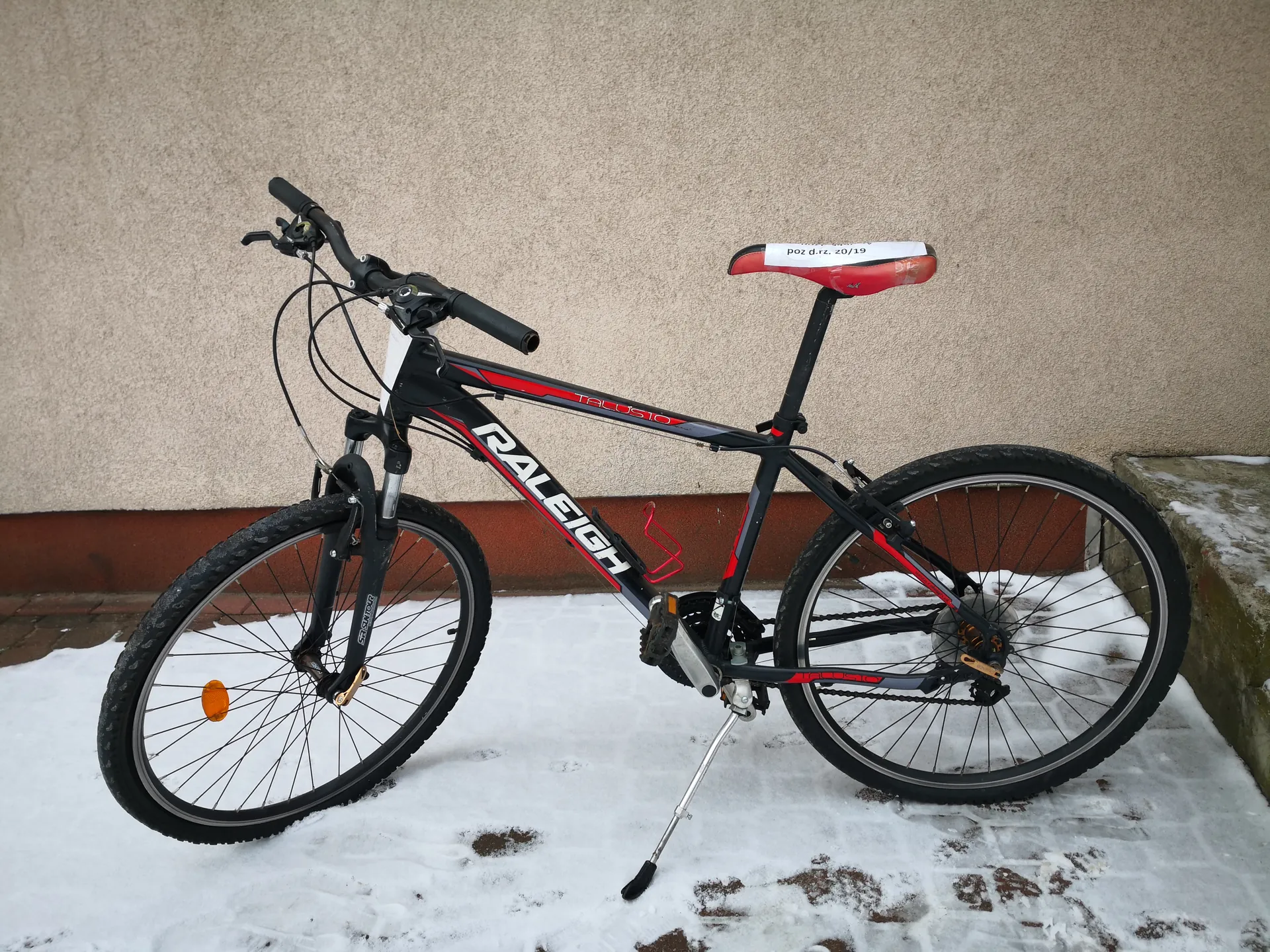 Czarny rower marki RALEIGH z czerwonymi akcentami, stojący na śniegu przed beżową ścianą z brązowym pasem u dołu. Na ramie widoczny napis 'TALUS 10', na siedzisku biała etykieta z tekstem 'poz d.rz. 20/19'. Rower ma czarne koła z srebrnymi obręczami, pomarańczowy reflektor na przednim kole i czerwony uchwyt na bidon. Tło stanowi tekstura ściany i śnieg na ziemi z widocznymi plamami ziemi.