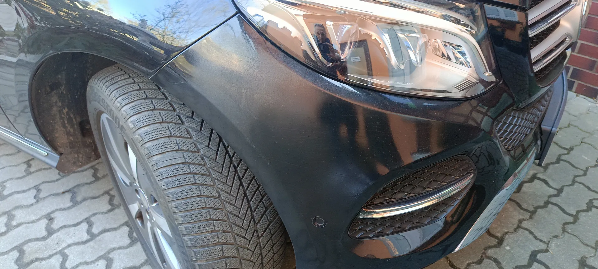 Czarny samochód widziany z przodu po lewej stronie, z widocznym reflektorem, częścią atrapy chłodnicy, zderzakiem i przednim lewym kołem z oponą o widocznym wzorze. Samochód zaparkowany na brukowanej powierzchni, po prawej stronie ceglana ściana. W reflektorze odbicie osoby robiącej zdjęcie oraz budynków.