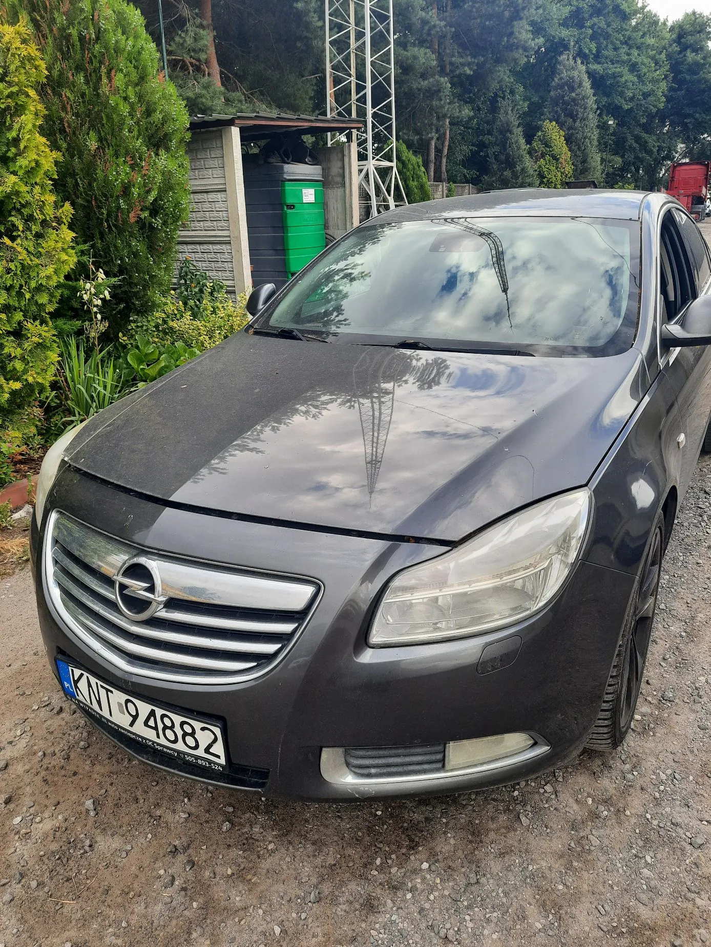 Zdjęcie przodu ciemnoszarego samochodu marki Opel. Pojazd posiada tablicę rejestracyjną o numerze KNT 94882. W tle widoczna jest zielona roślinność, krzewy, mały budynek gospodarczy z zielonym zbiornikiem oraz metalowa konstrukcja wieży.