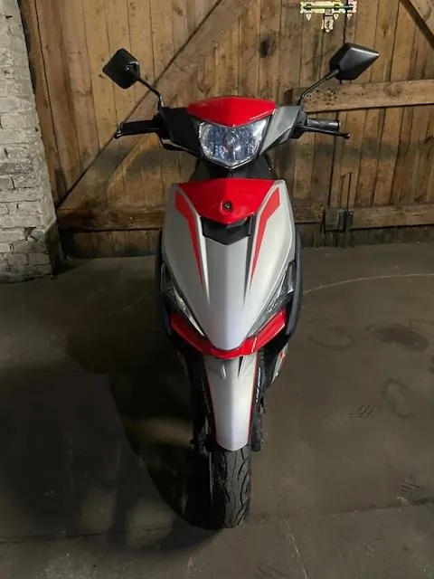 Czerwono-biały skuter widziany od przodu, z reflektorem, lusterkami i czarnymi elementami. Skuter stoi na betonowej powierzchni przed drewnianą bramą z metalowymi zawiasami. Po lewej stronie widoczna jest ceglana ściana. Na podłodze znajdują się plamy i ślady.