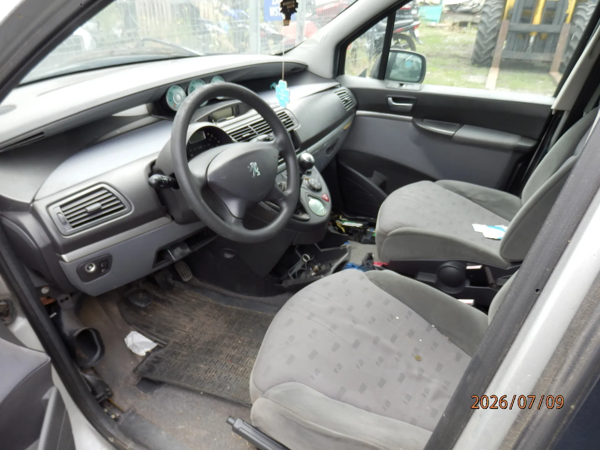 Wnętrze samochodu widziane z perspektywy otwartych drzwi pasażera. Widoczna jest szara deska rozdzielcza, kierownica z logo marki Peugeot, dwa przednie szare fotele materiałowe oraz gumowe dywaniki na podłodze. W prawym dolnym rogu znajduje się data 2026/07/09.