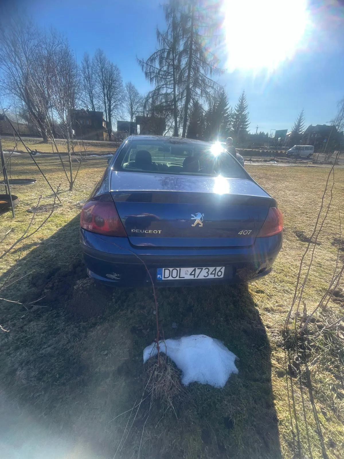 Z tyłu niebieski samochód Peugeot 407 z tablicą rejestracyjną DOL 47346 (PL po lewej stronie). Na bagażniku widoczny logo lwa i napis PEUGEOT. Samochód stoi na trawie z resztkami śniegu, w tle drzewa bez liści, domy, jasne niebo i słońce oświetlające scenę. Po prawej stronie widoczna postać człowieka oraz biały samochód w oddali.