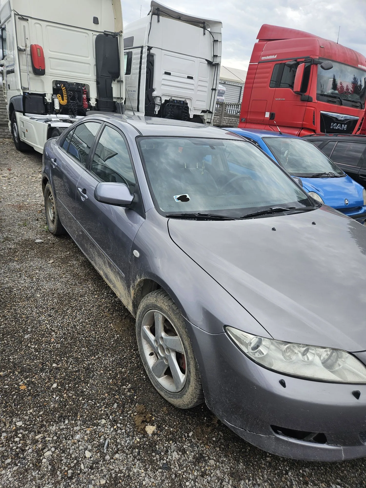Szarosrebrny samochód osobowy typu sedan zaparkowany na żwirowym placu. Auto jest zabrudzone błotem w dolnych partiach nadwozia. W tle widoczne są duże pojazdy ciężarowe: biała naczepa oraz czerwona ciężarówka marki MAN, a obok szarego sedana stoi niebieski samochód osobowy.