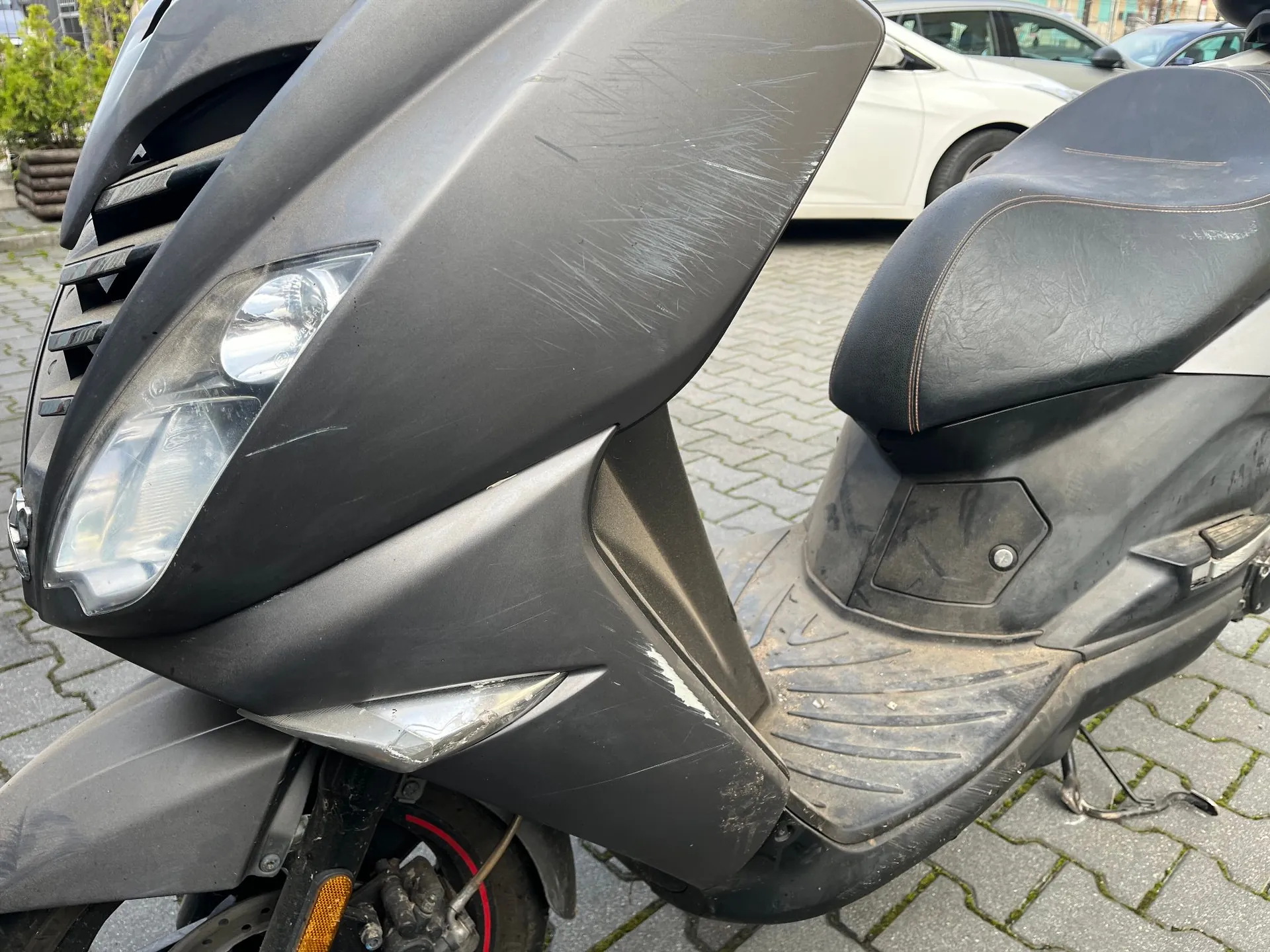 Zbliżenie na przednią część czarnego skutera zaparkowanego na kostce brukowej. Na plastikowej obudowie skutera, w okolicach reflektora, widoczne są liczne zarysowania i przetarcia. W tle widać fragment białego samochodu.