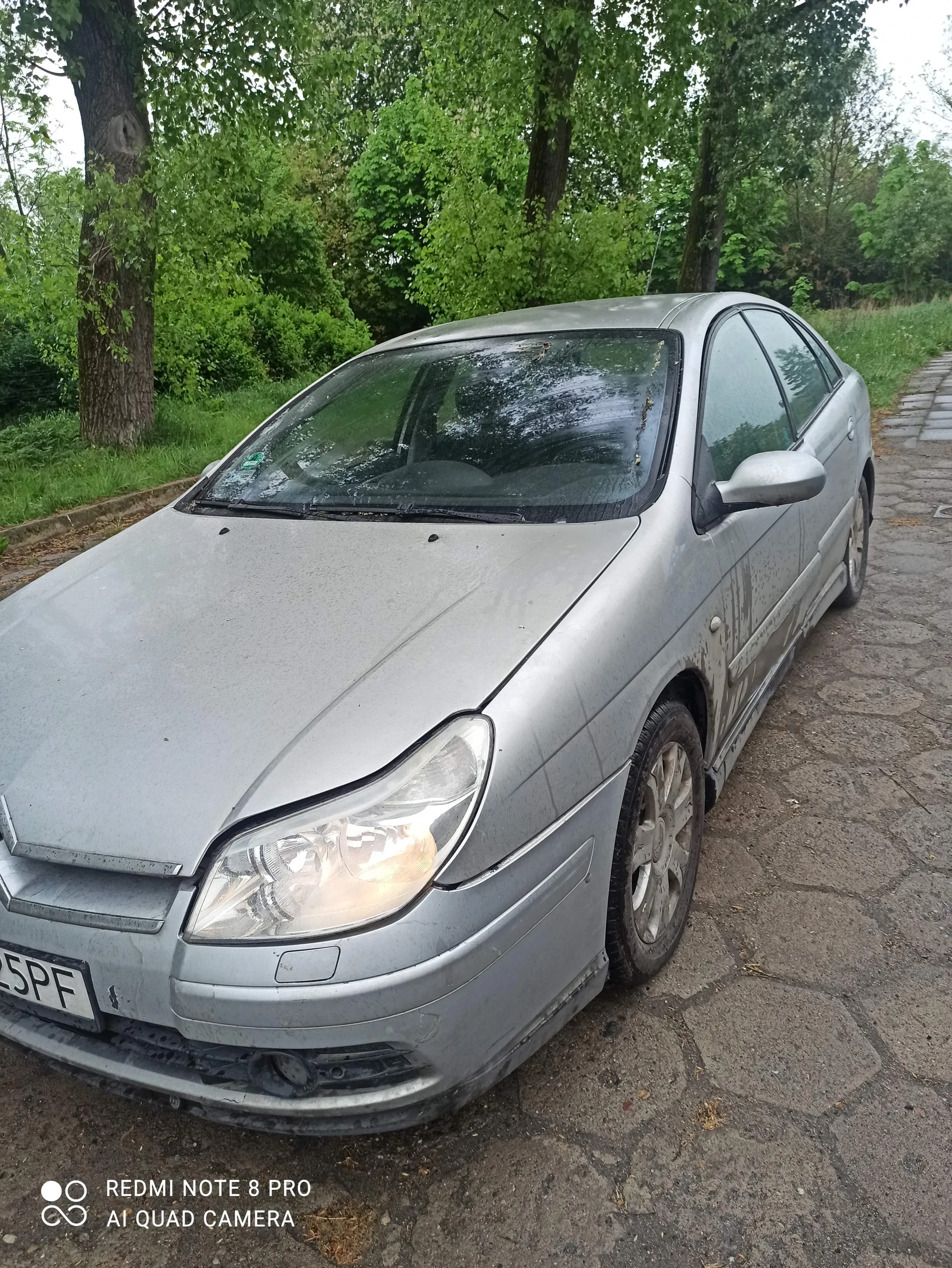 Srebrny samochód zaparkowany na brukowanej powierzchni. Widoczne są zanieczyszczone karoseria, przednie światło, koła oraz częściowo czytelna tablica rejestracyjna z napisem 5PF. W tle zielone drzewa i trawa. Po prawej stronie obrazu widoczny pionowy napis: REDMI NOTE 8 PRO AI QUAD CAMERA.