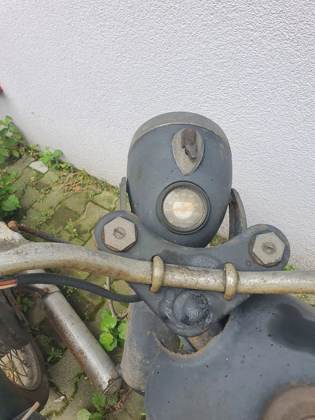 Zbliżenie na kierownicę i licznik starego motocykla. Widoczny jest okrągły wskaźnik prędkości z cyframi od 0 do 100, zamontowany w ciemnoszarej obudowie. Kierownica jest metalowa, z widocznymi śladami korozji i rdzy. Motocykl stoi na kostce brukowej porośniętej mchem i trawą, obok białej, chropowatej ściany.